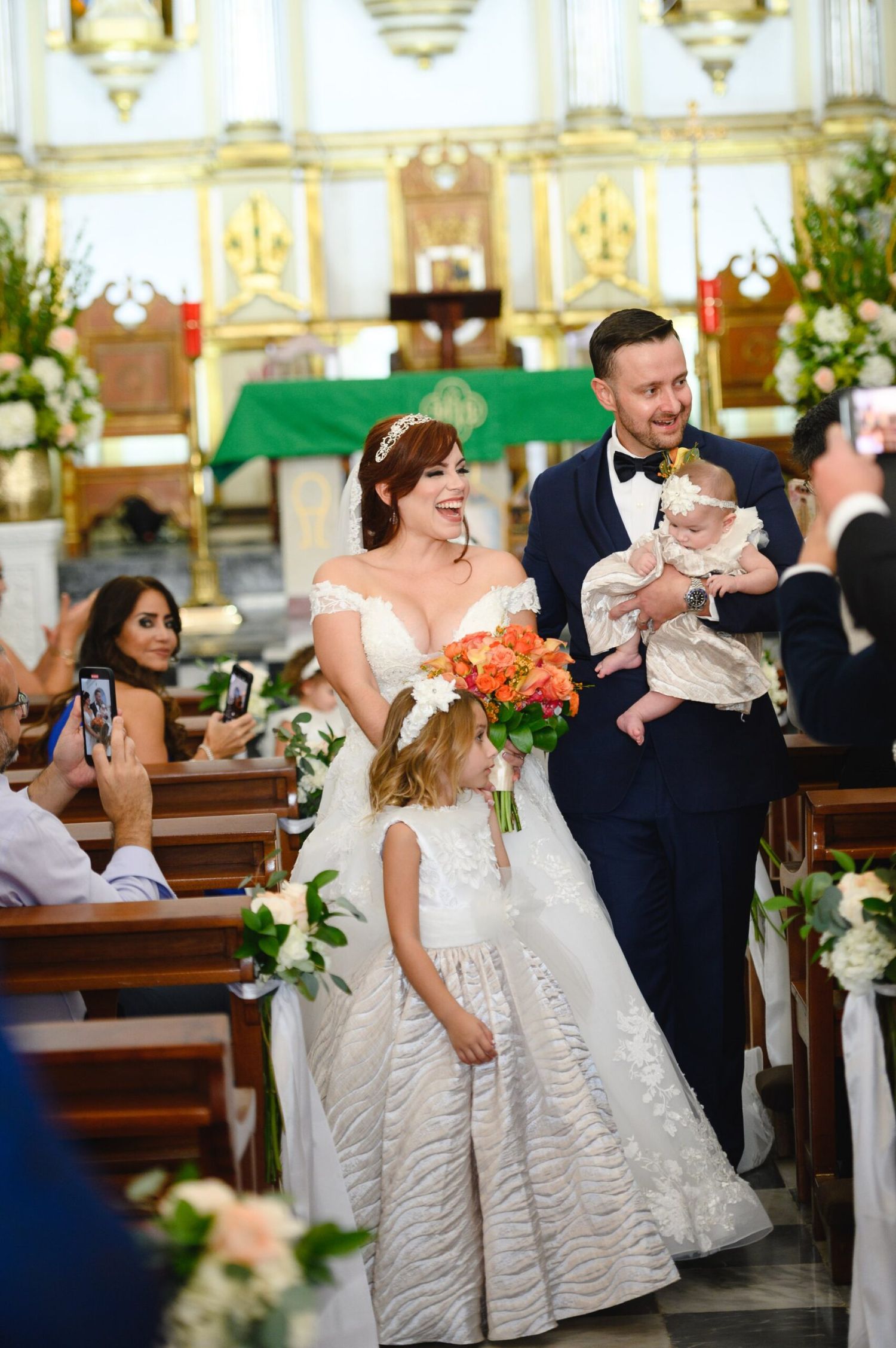 Bride and groom walking down church aisle with flower girl and baby during wedding ceremony.