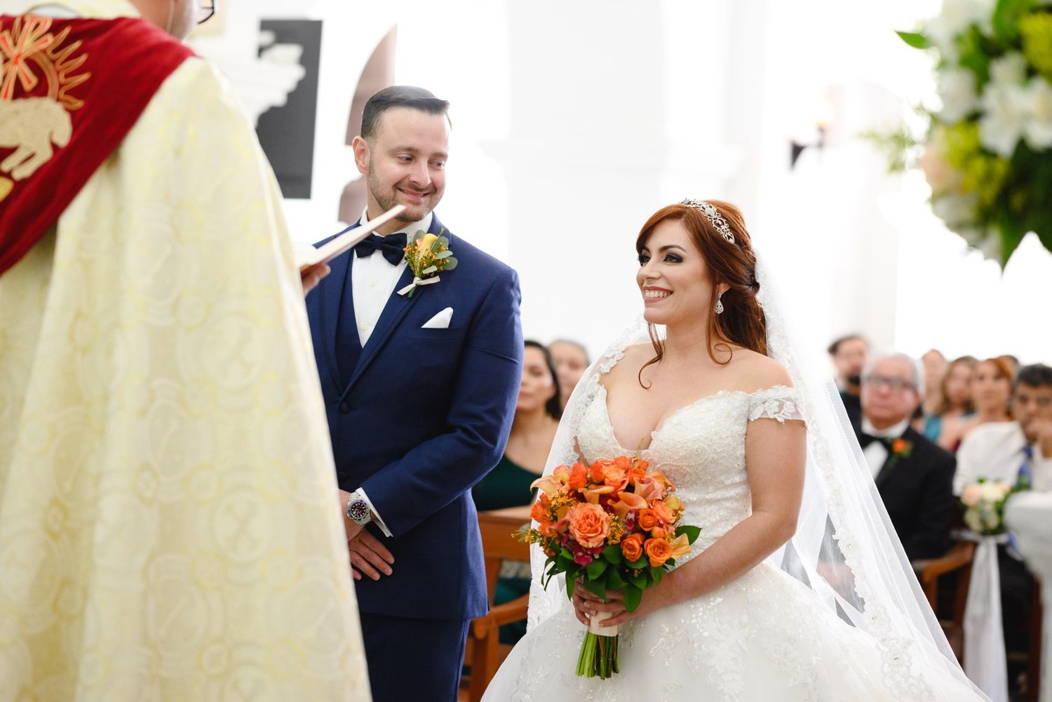 Bride holding orange bouquet smiles at groom in blue suit during church wedding ceremony.