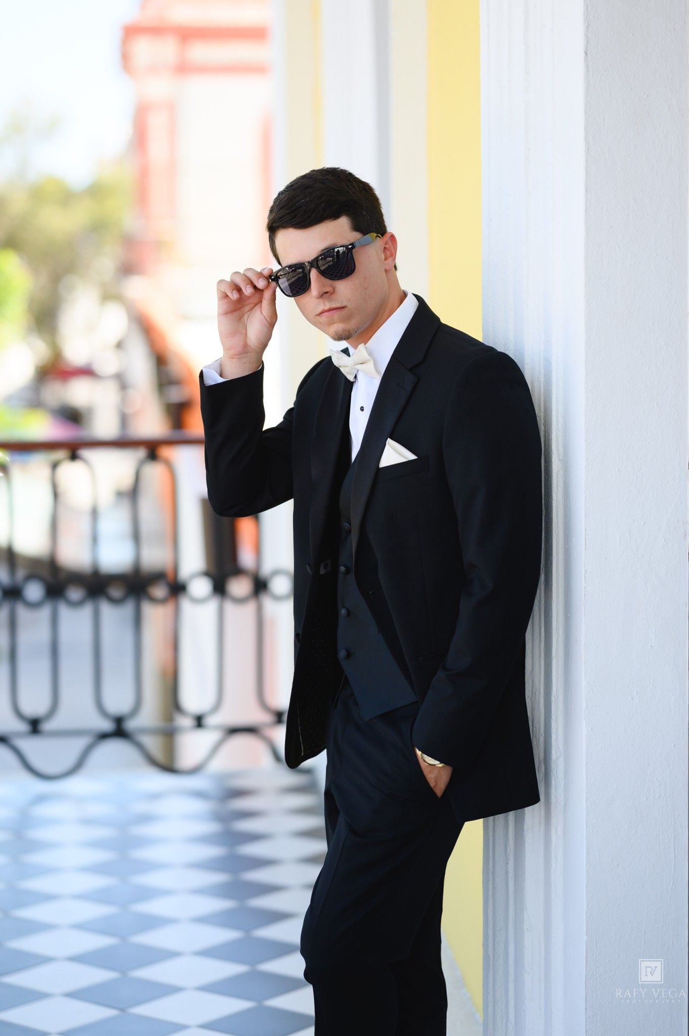 Young man in black tuxedo and sunglasses leaning against white column outdoors.