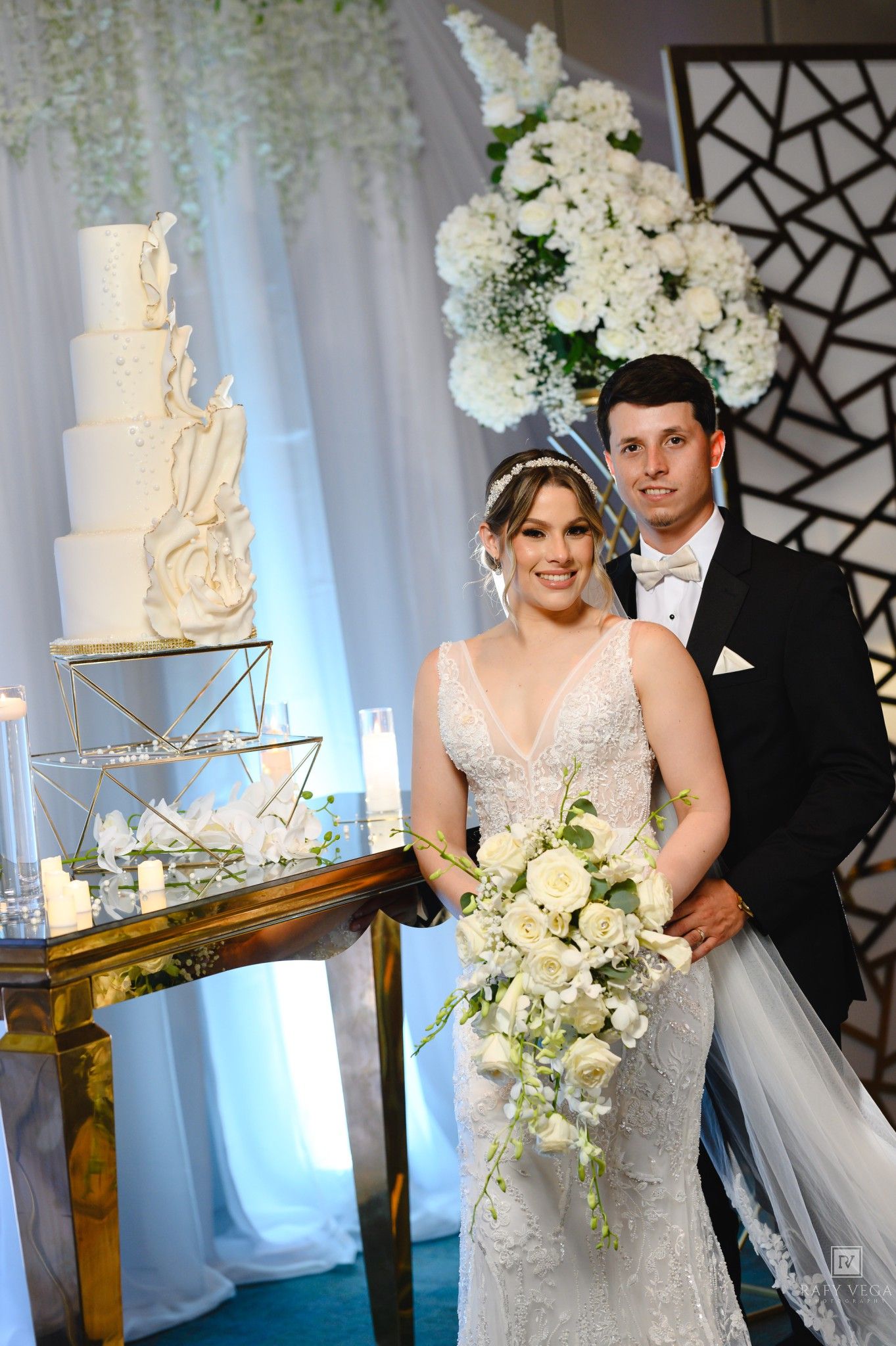Bride and groom pose beside elegant white wedding cake with cascading floral bouquet at reception.