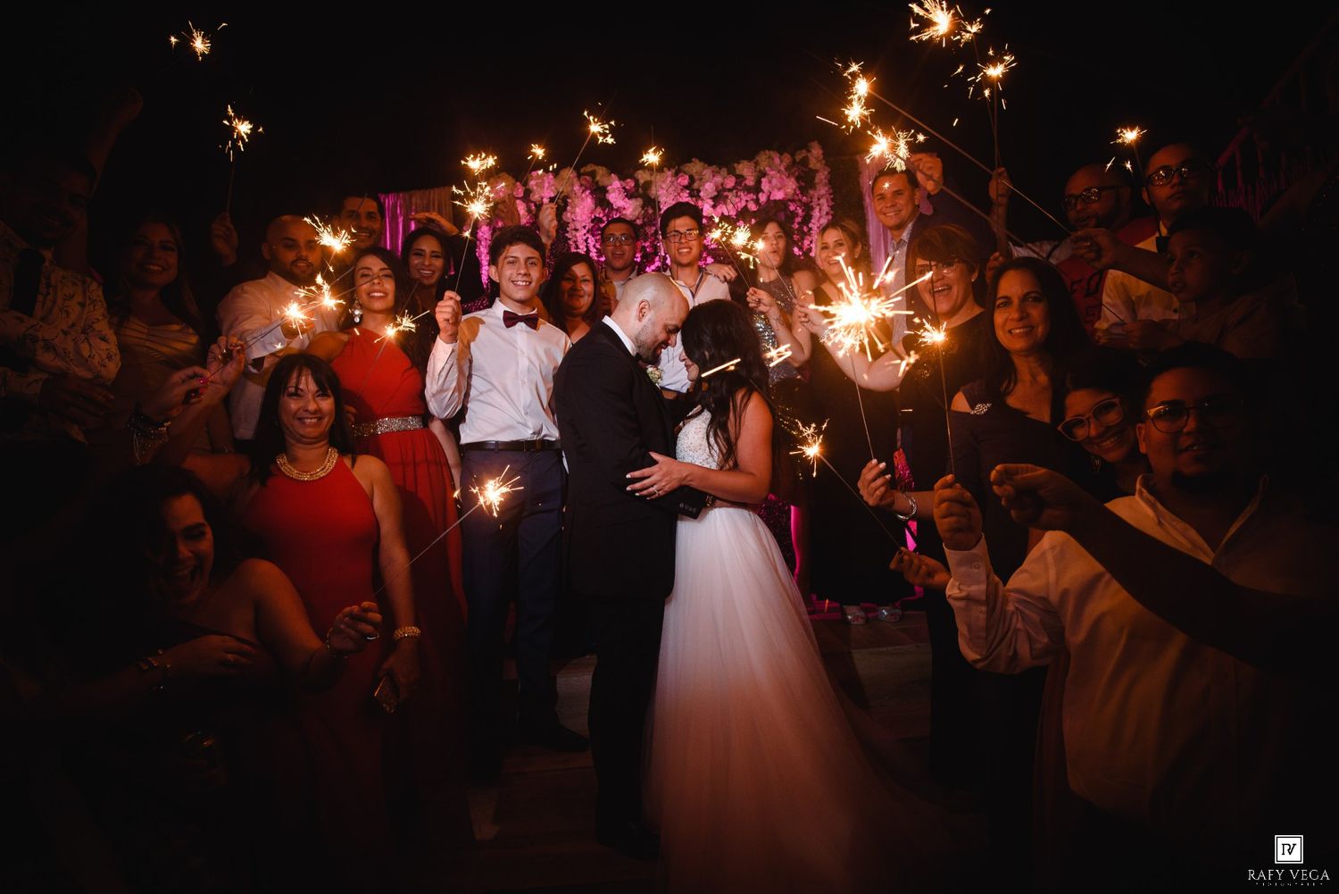 Bride and groom share a romantic kiss surrounded by guests holding sparklers at night.
