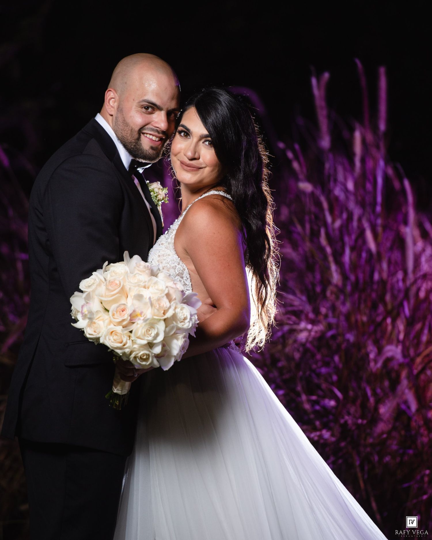 Smiling bride and groom pose together at night with purple ornamental grasses, bride holding white rose bouquet.