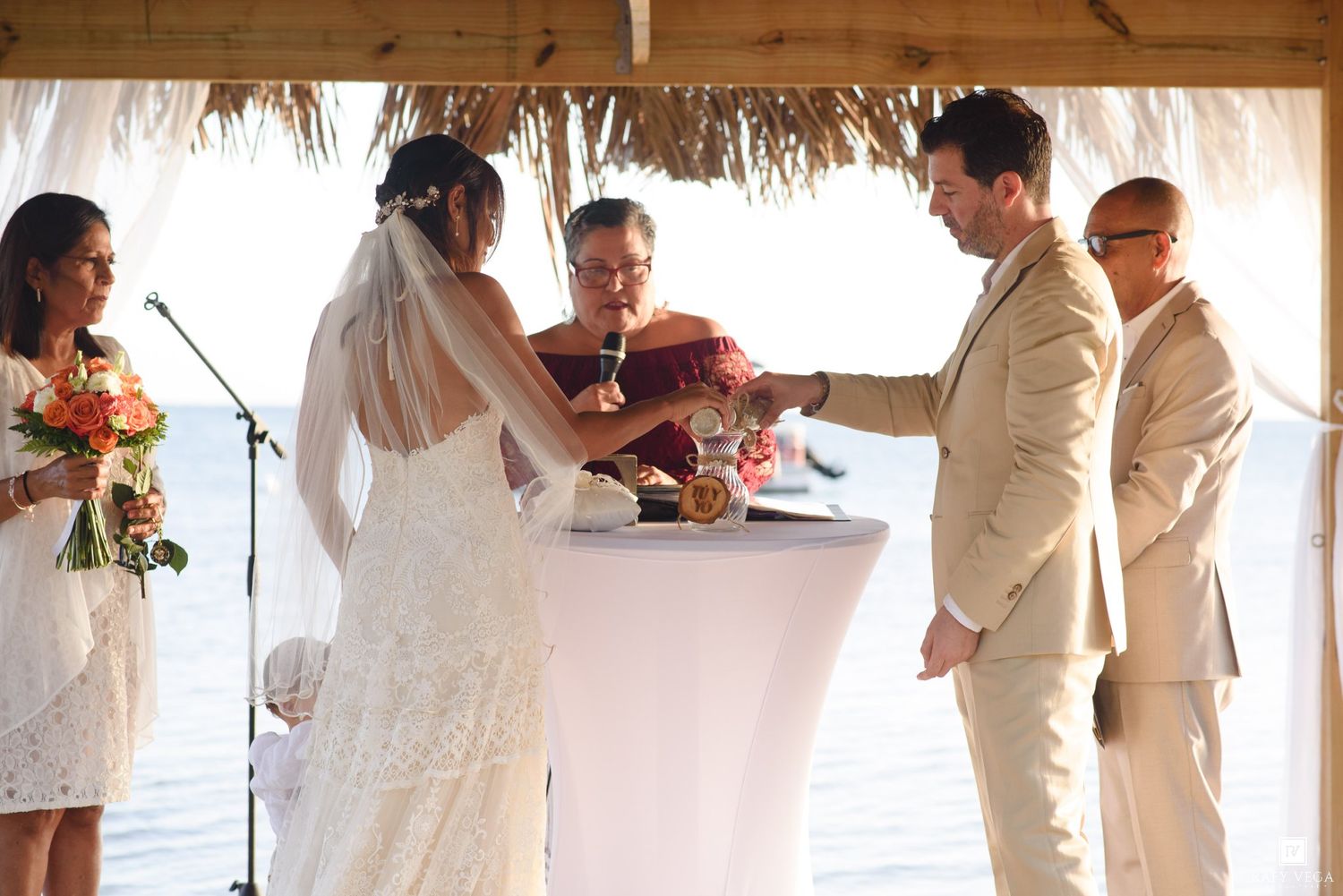 Bride and groom exchange vows at a beachside wedding ceremony under a thatched canopy with officiant and guests.
