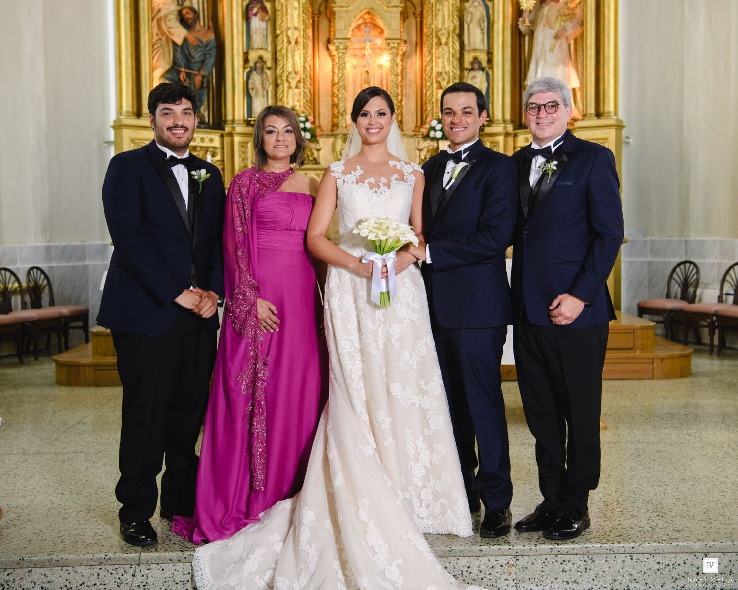 Bride in white gown poses with family in church after wedding ceremony, ornate golden altar in background.