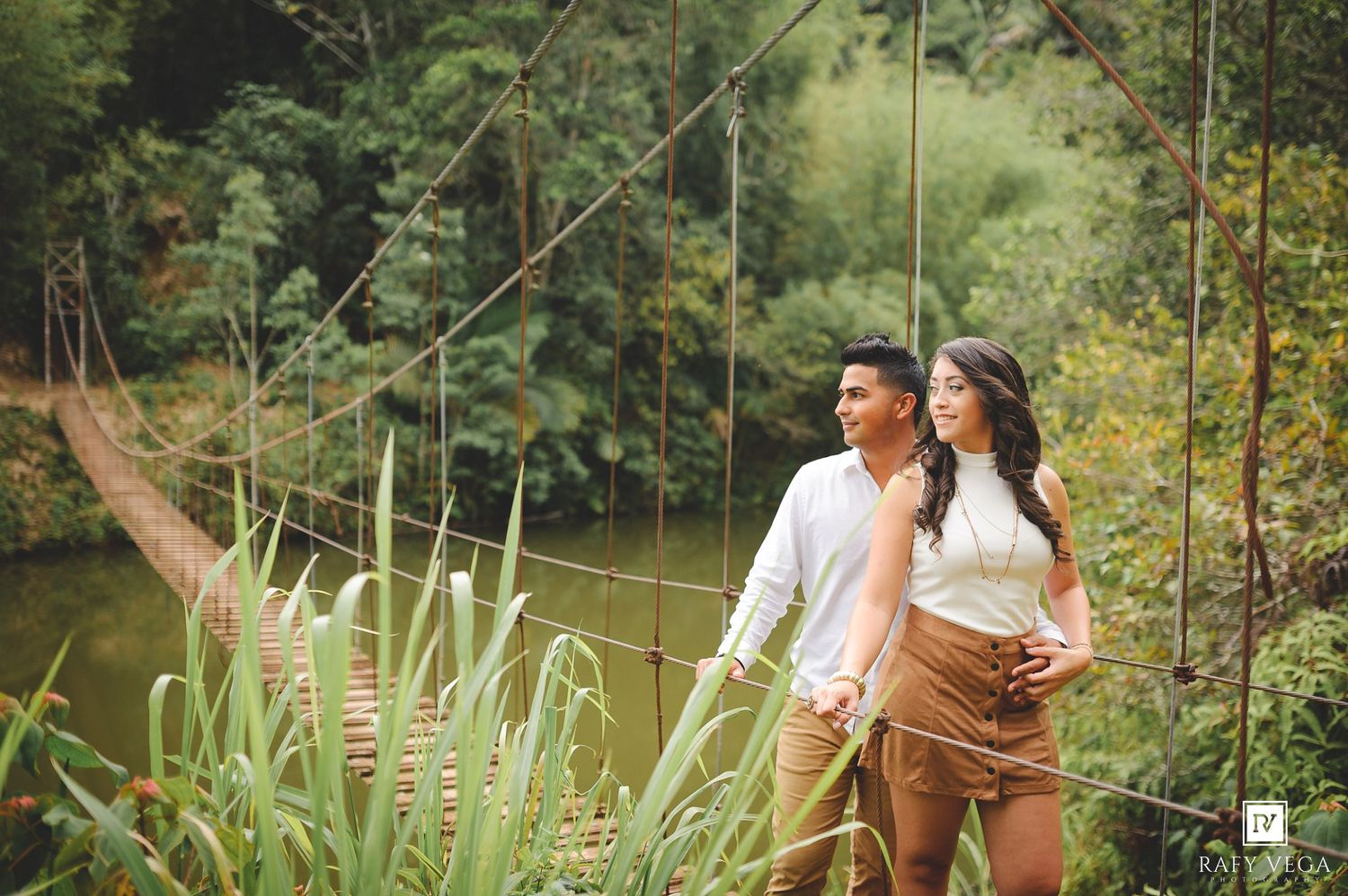 Couple holding hands on a rustic suspension bridge surrounded by lush green trees and a serene river backdrop.