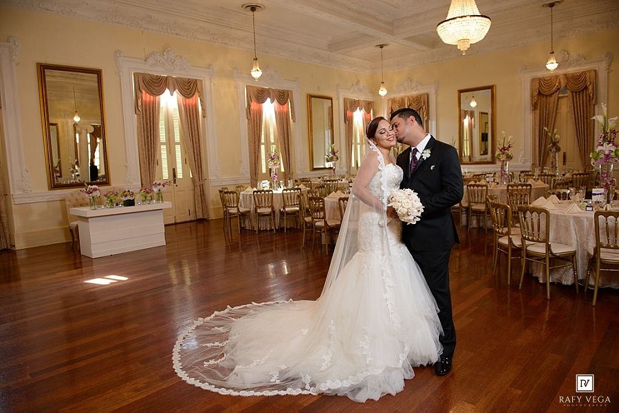 Bride and groom share a romantic embrace on a polished dance floor in an elegant gold-accented wedding reception hall.
