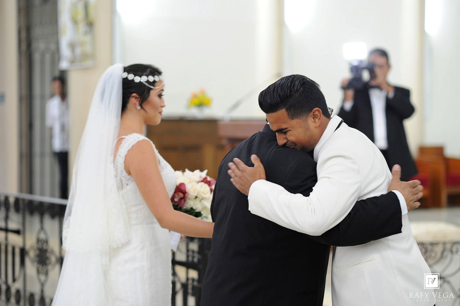 Bride in white gown and groom in white suit share an emotional hug with a guest during a church wedding ceremony.