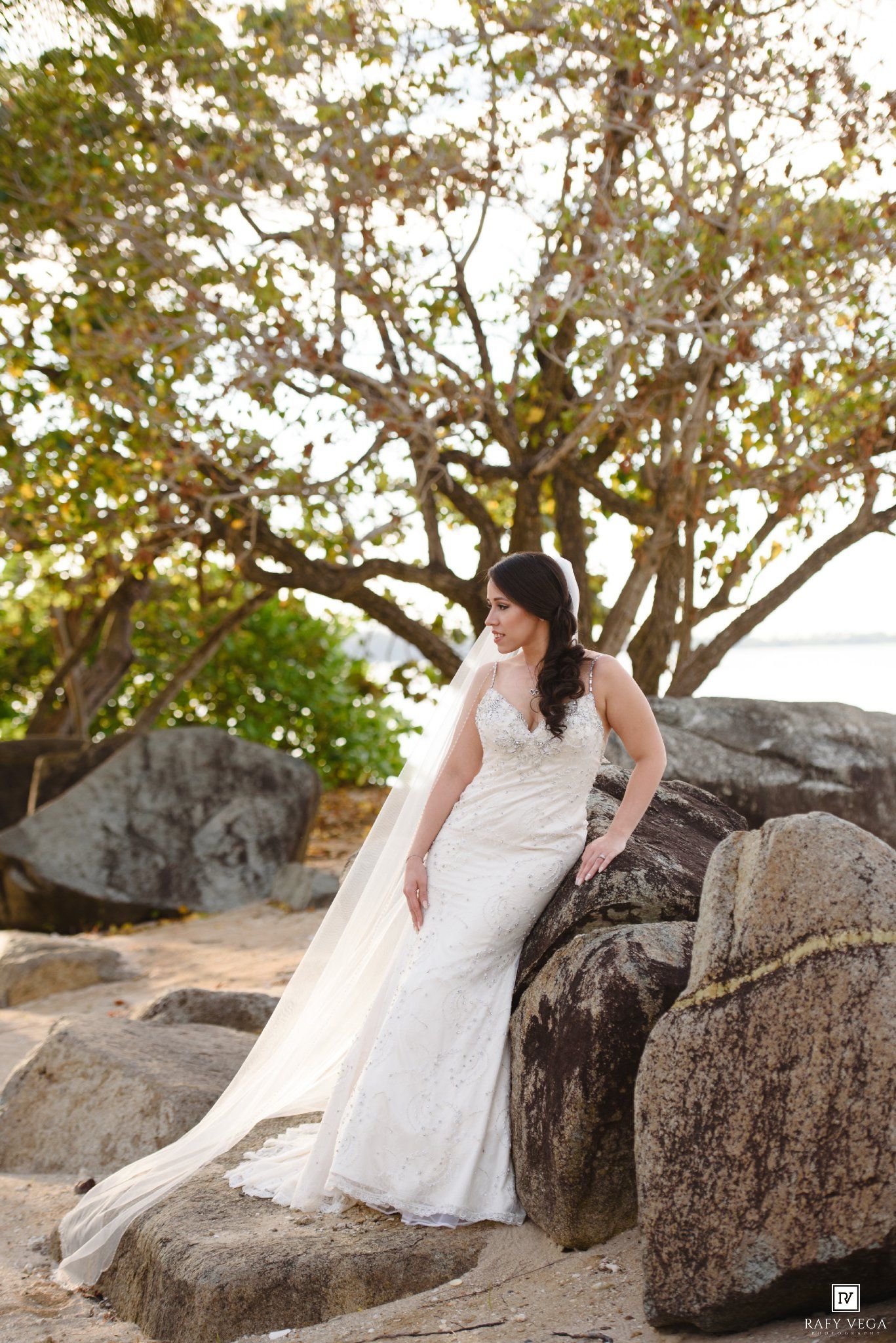 Bride in white lace wedding gown and veil posing on rocks near beach with trees in background.