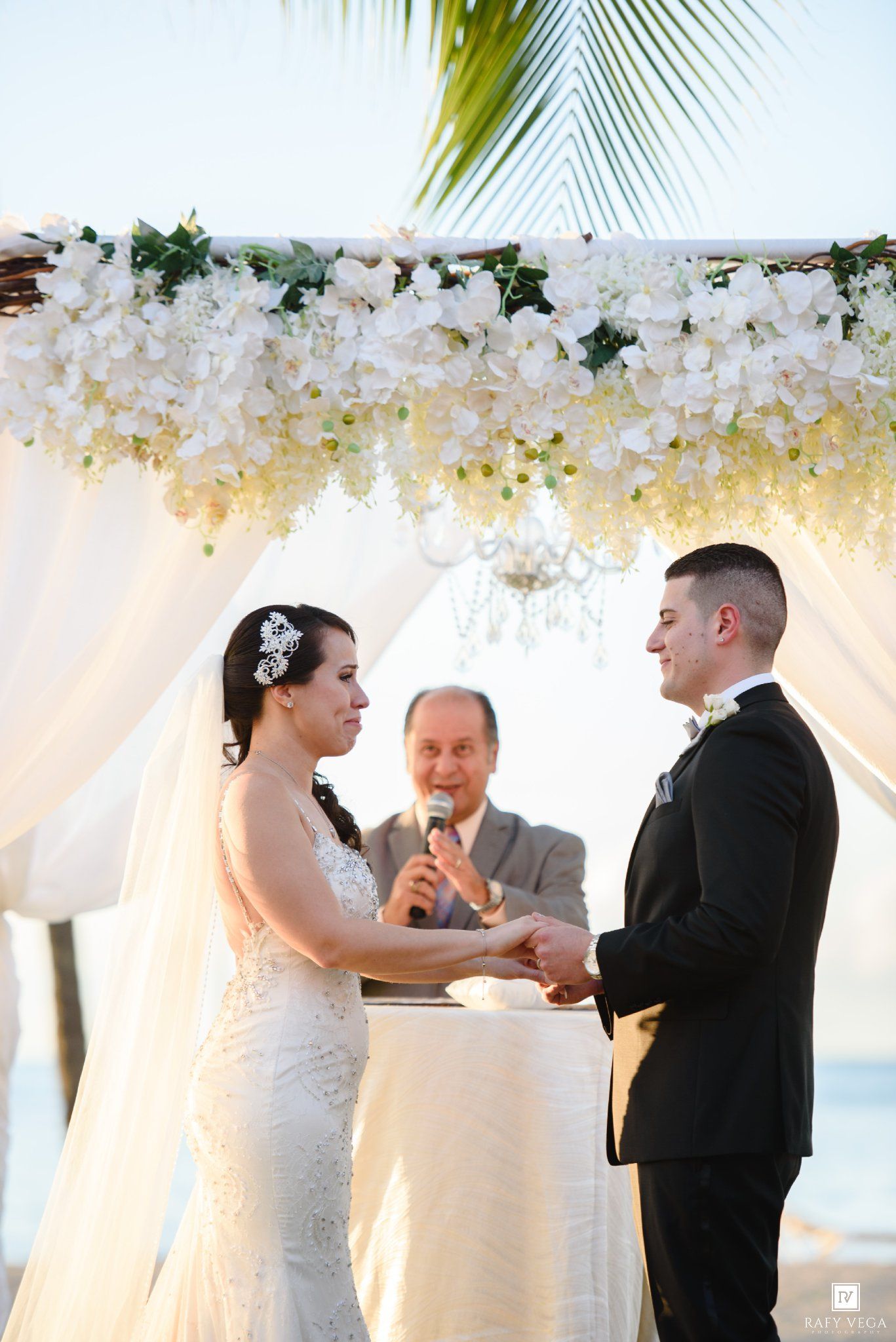 Bride and groom exchange vows under a white floral arch at a beach wedding ceremony with an officiant.