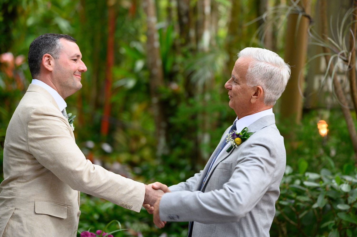 Two men in suits shaking hands at an outdoor wedding ceremony surrounded by lush greenery.