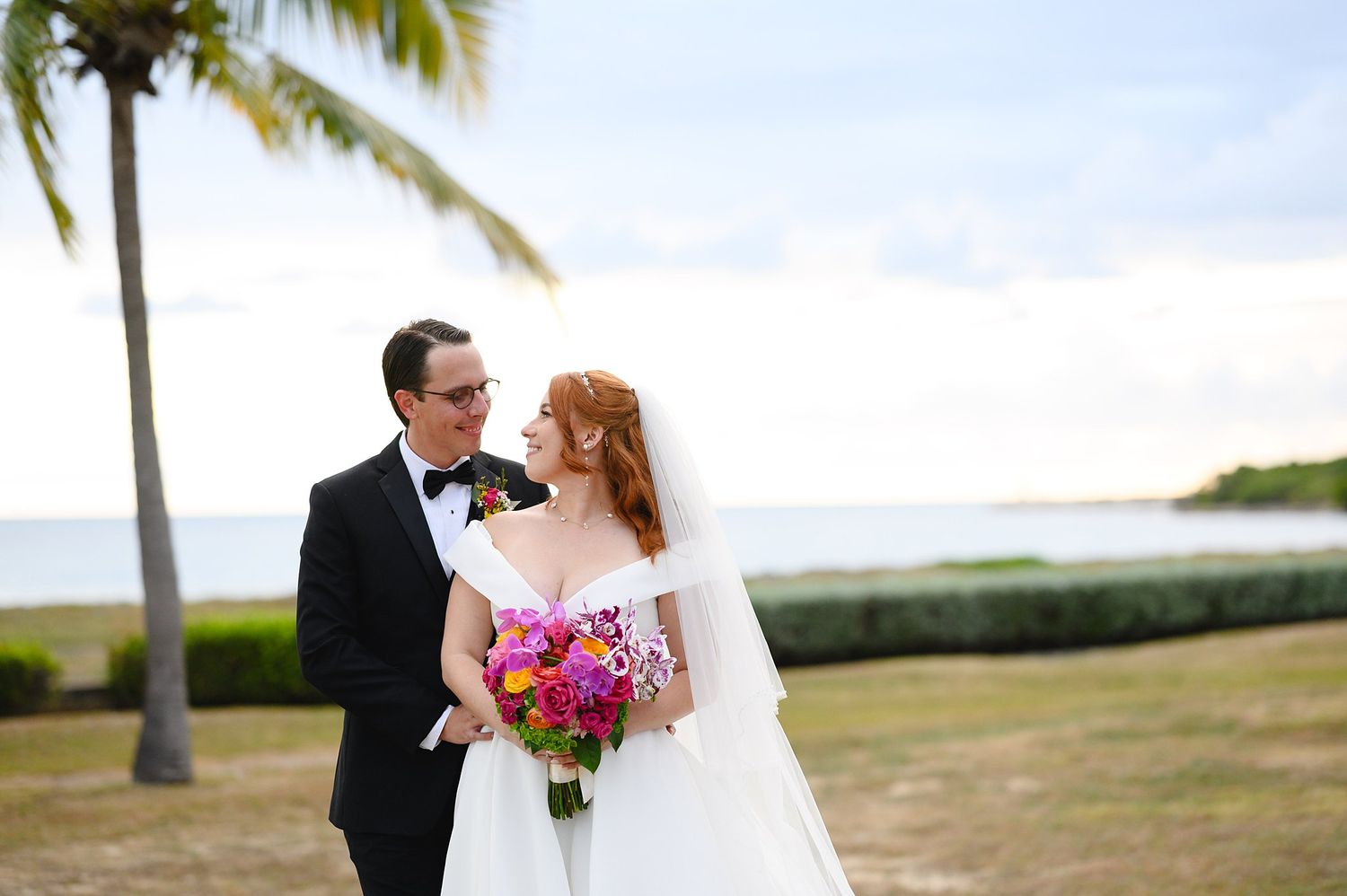 Bride and groom share a romantic moment outdoors near a palm tree and ocean, bride holding vibrant pink and purple bouquet.