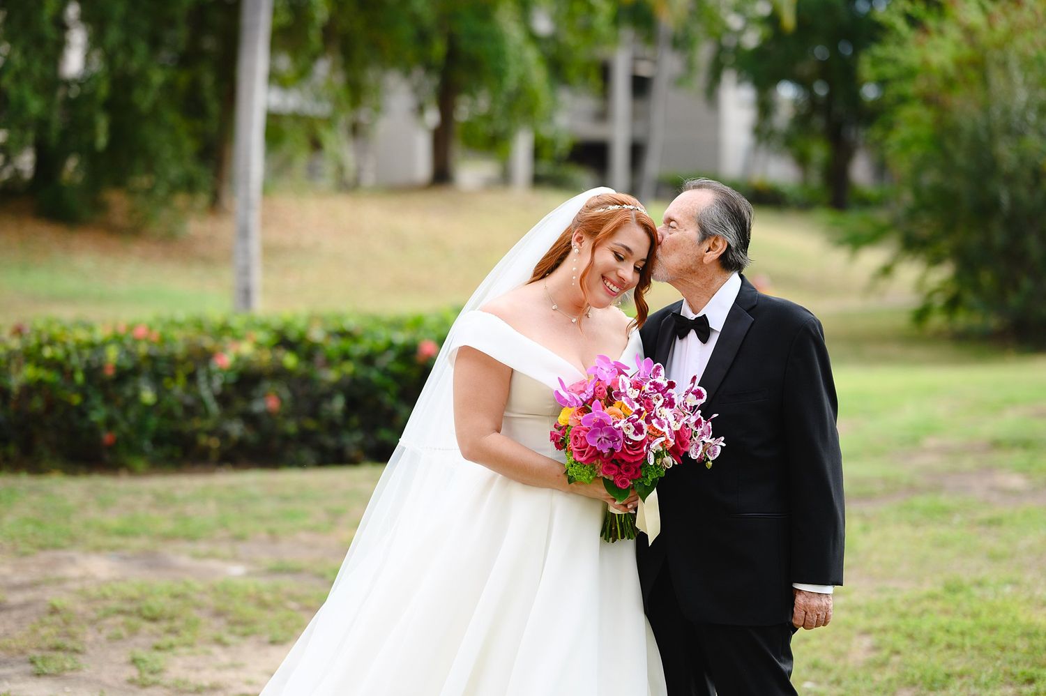 Bride in white off-shoulder gown holds pink bouquet beside man in tuxedo outdoors near lush greenery.