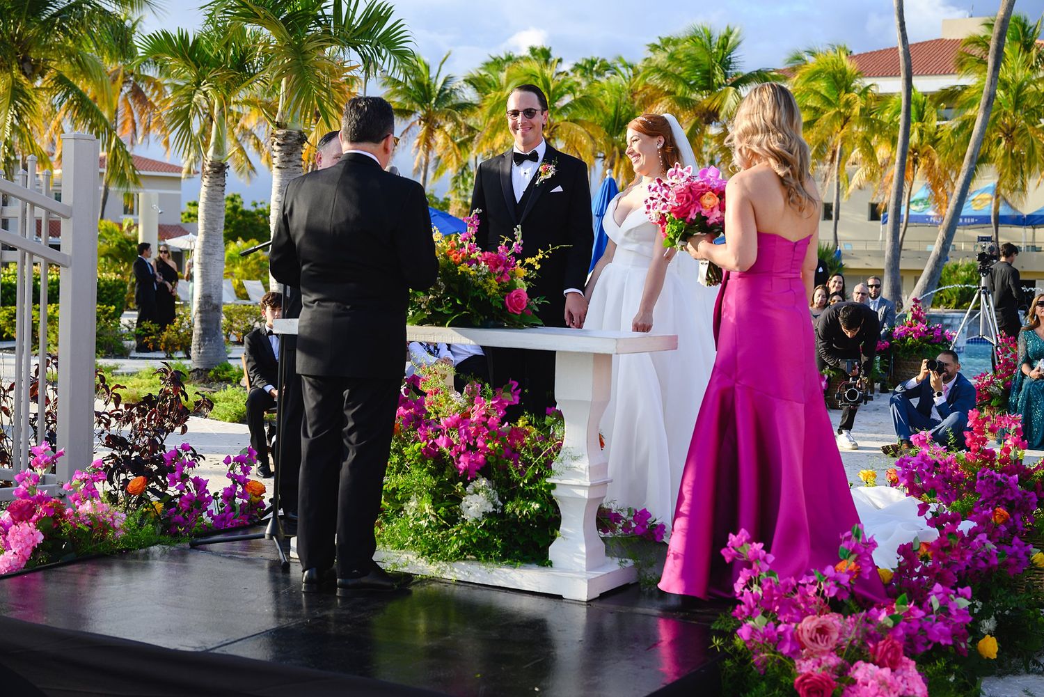 Outdoor wedding ceremony with bride and groom at floral-decorated altar, surrounded by colorful blooms and palm trees.