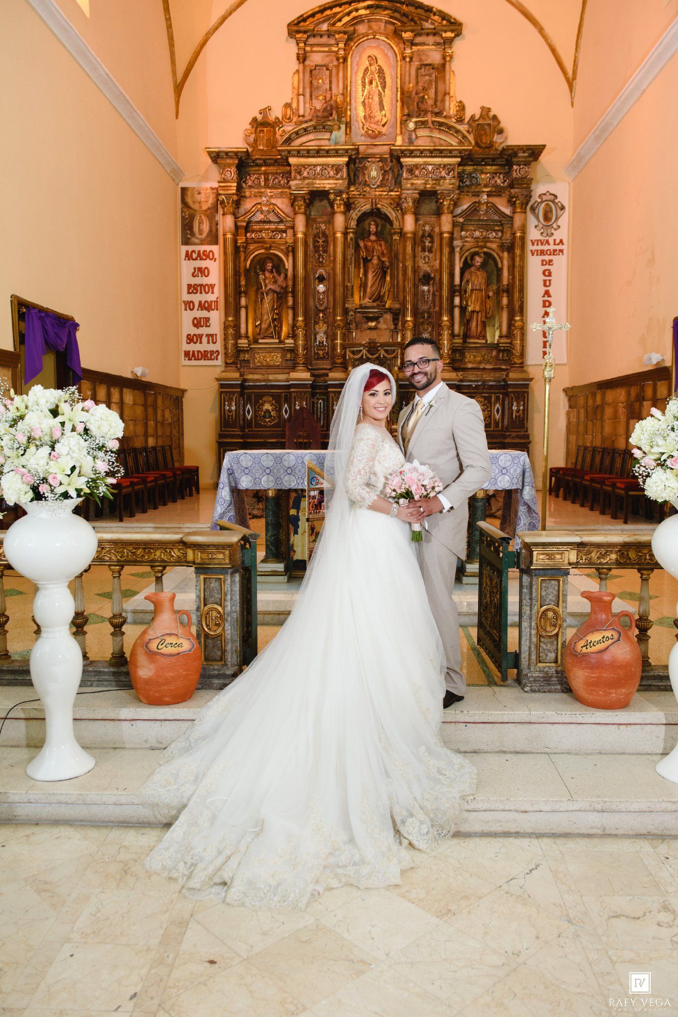 Bride and groom pose together at ornate church altar with golden decor and white floral arrangements on their wedding day.