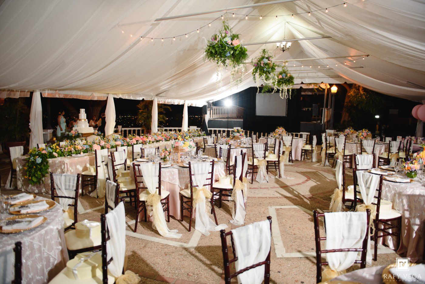 Elegant wedding reception under a white tent with chiavari chairs, floral centerpieces, and a tiered cake.