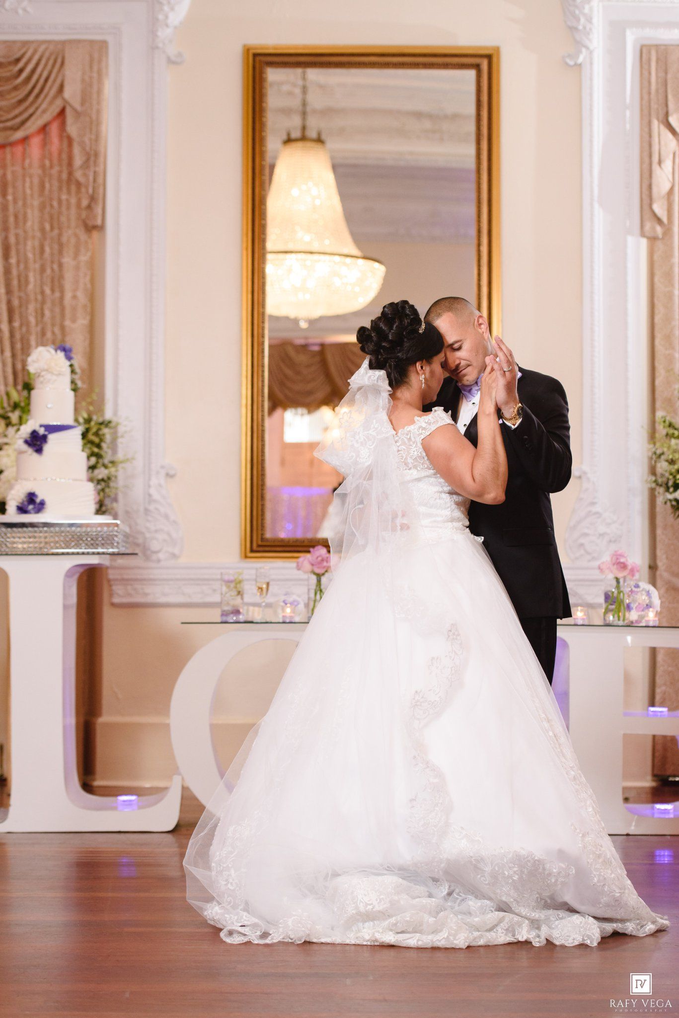 Bride and groom share their first dance in an elegant white wedding reception hall with a tiered cake nearby.
