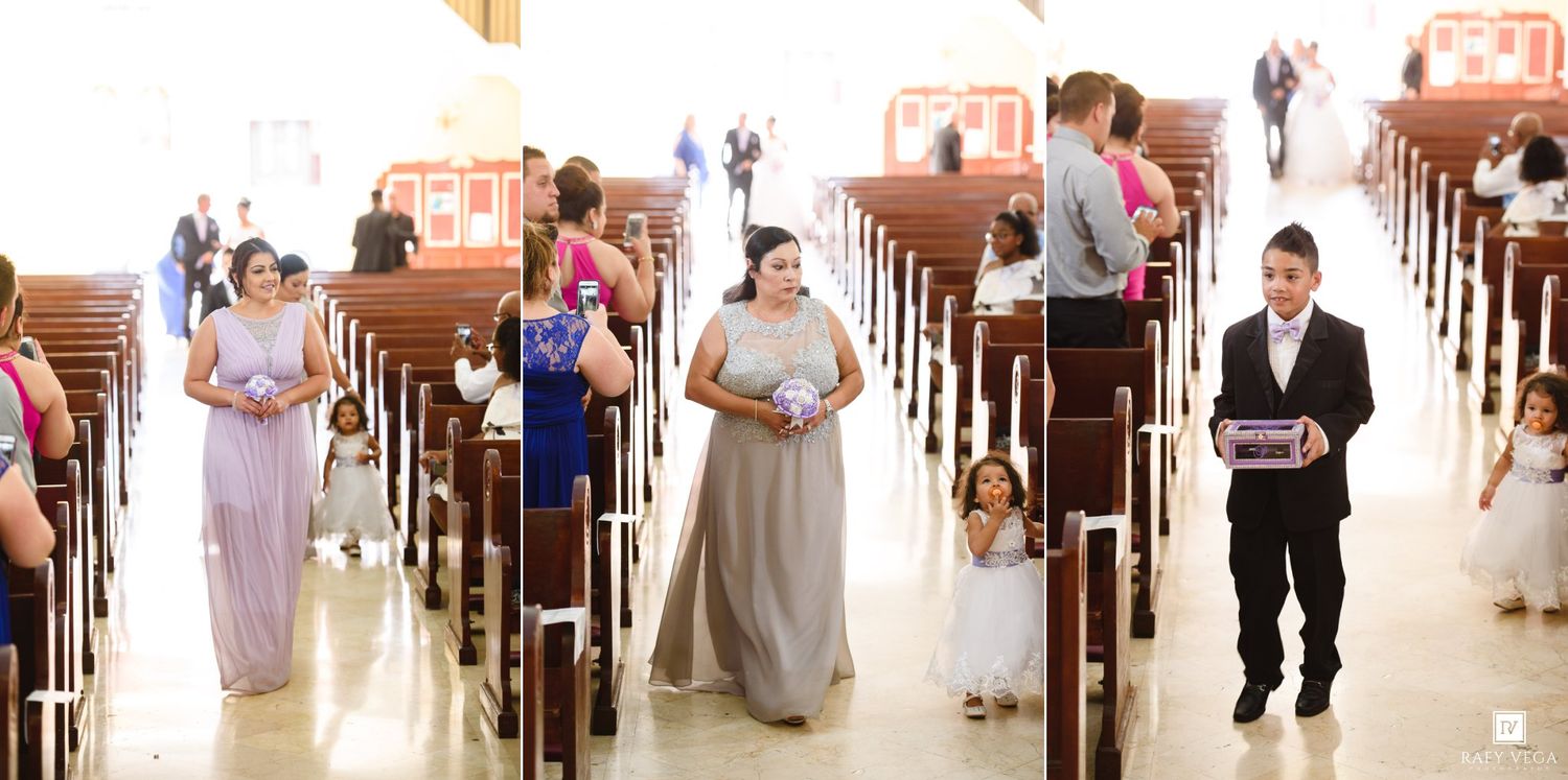 Bridesmaids in gray and blue gowns walk down a church aisle during a wedding ceremony with guests seated in pews.