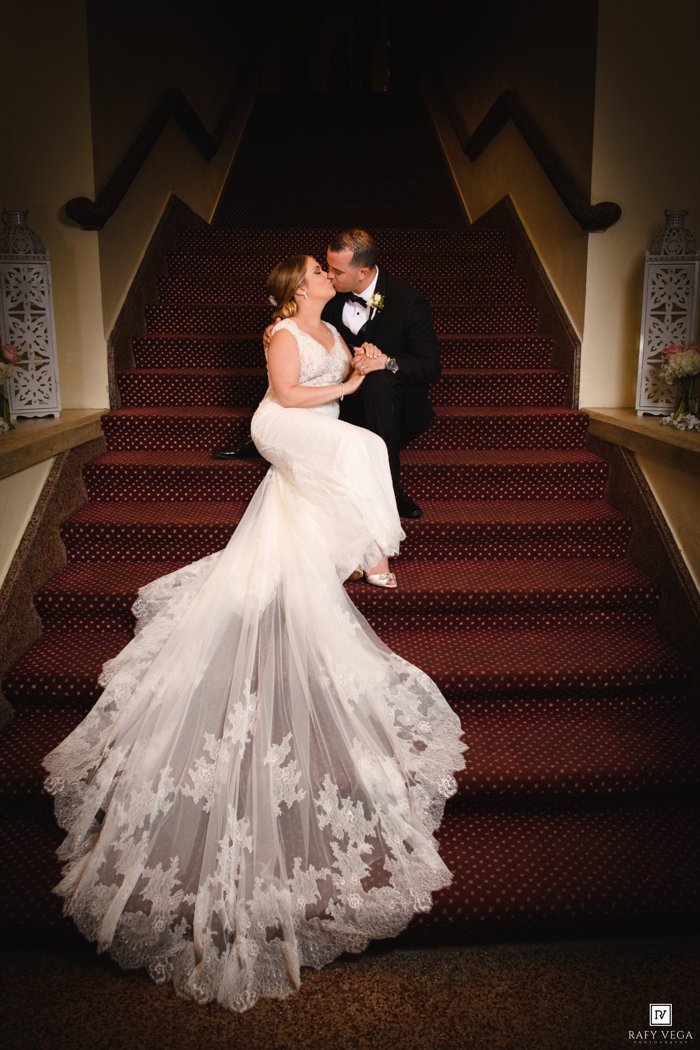 Bride and groom share a romantic kiss on red-carpeted stairs, her lace mermaid gown trailing elegantly below.