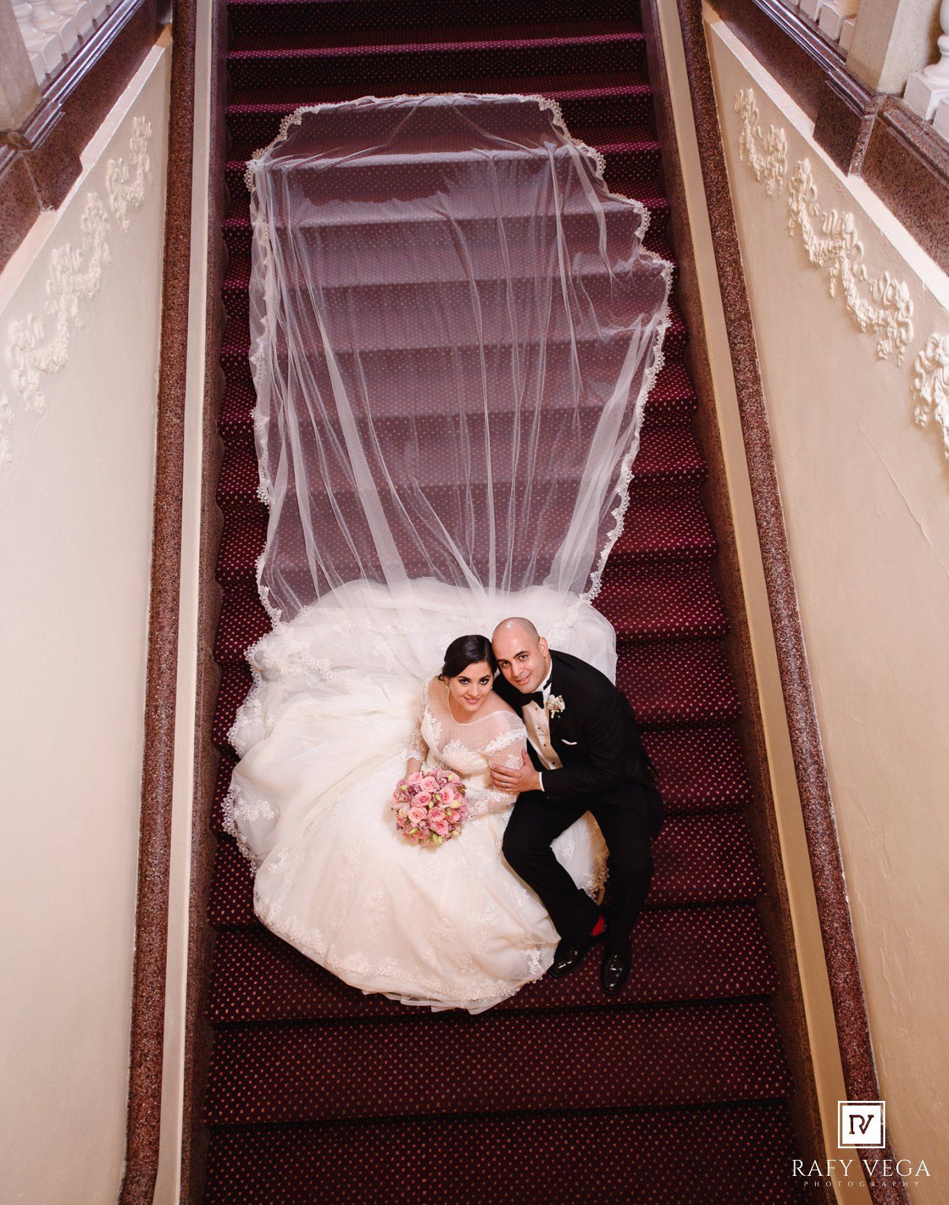 Aerial view of bride and groom sitting on red carpeted staircase with flowing white veil spread above.