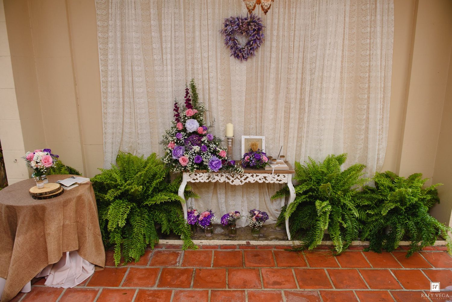 Elegant wedding display with purple floral arrangements, ferns, and a wreath on a white console table against a draped backdrop.