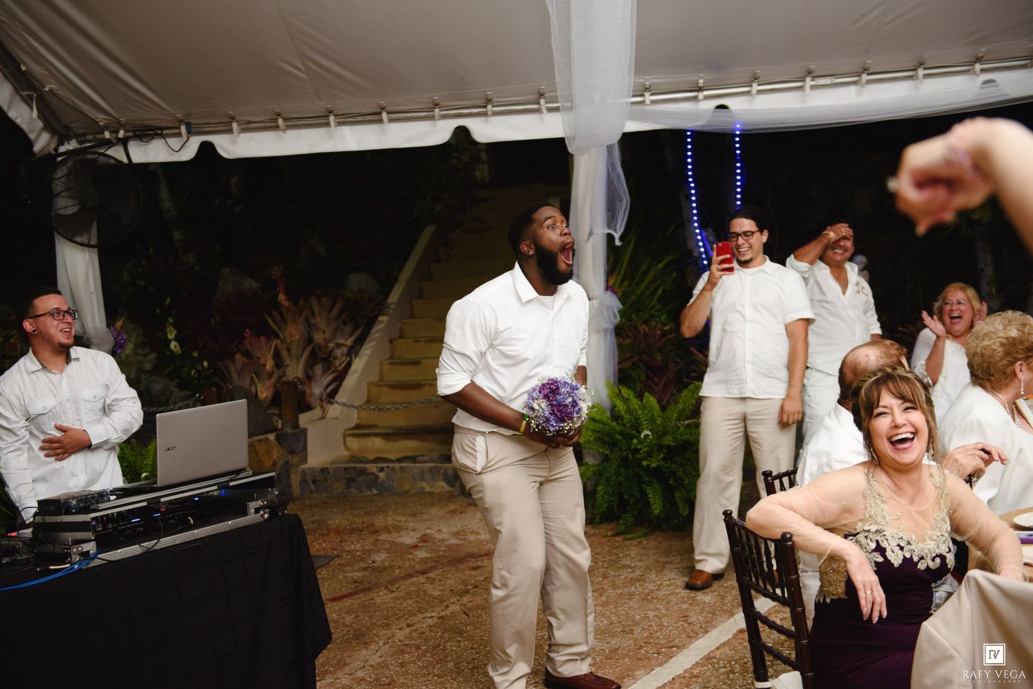 Groom in white suit holds purple bouquet at lively outdoor wedding reception under a tent at night.