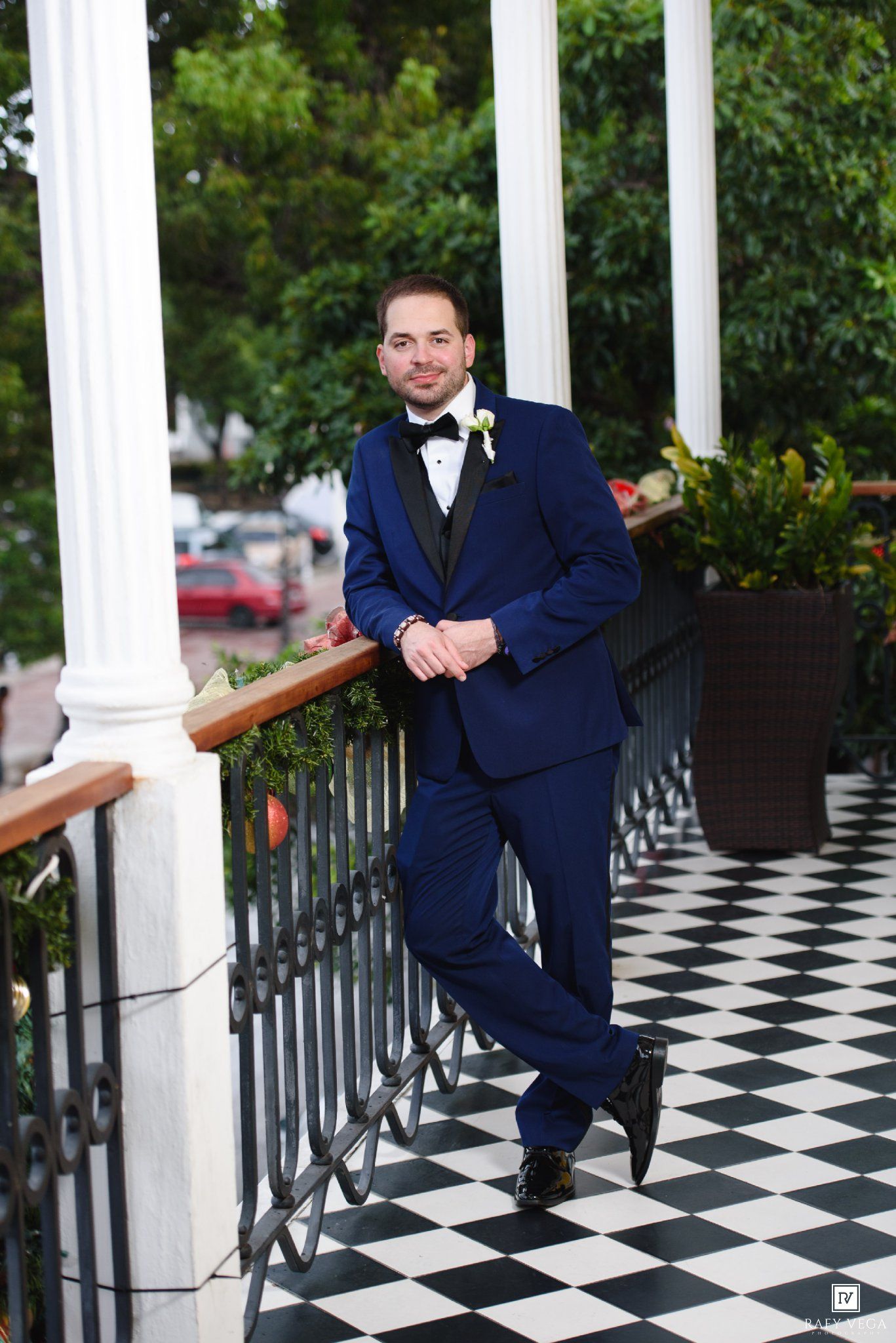 Groom in navy blue tuxedo posing on checkered floor balcony with white columns and greenery backdrop.