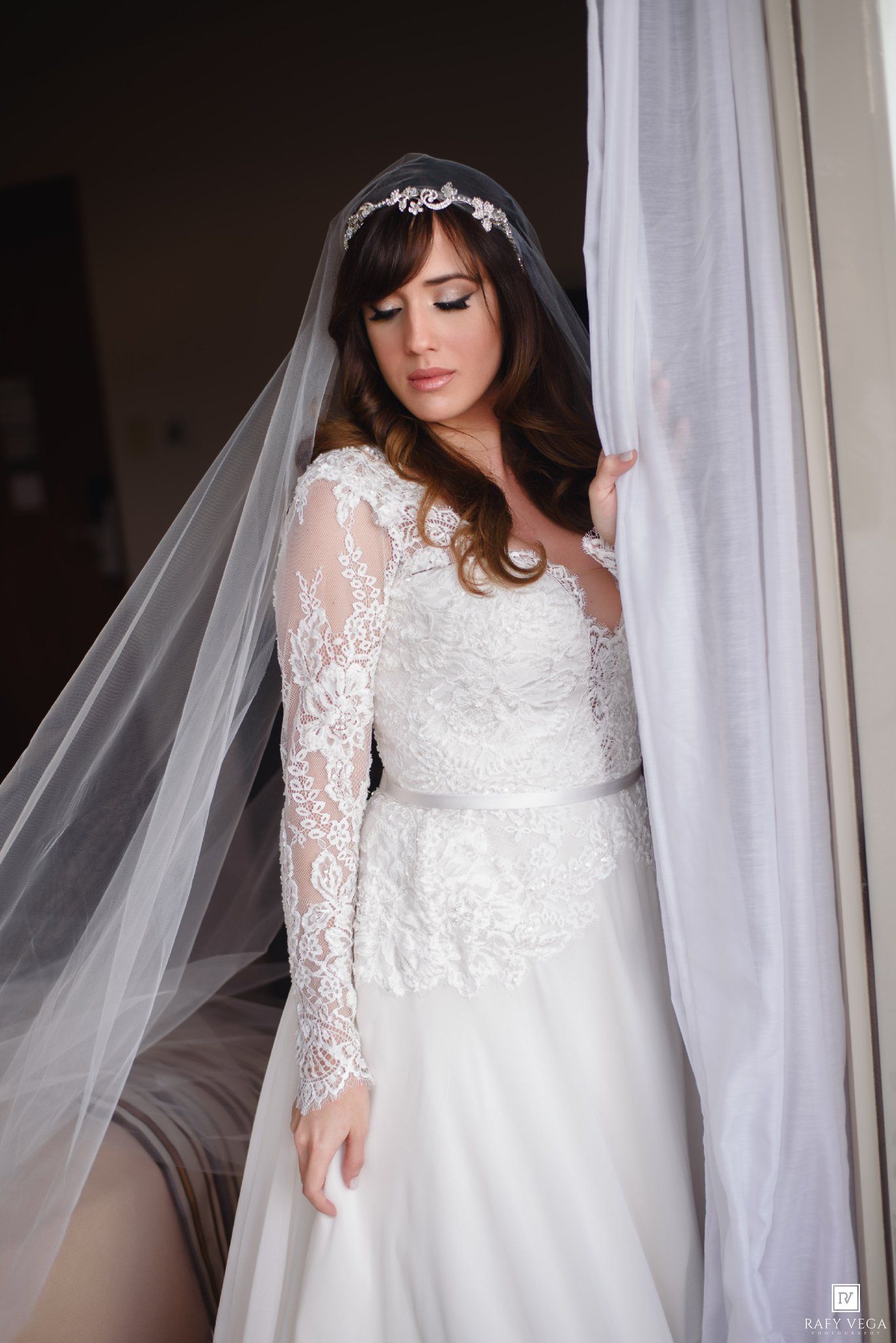 Bride in lace long-sleeve wedding gown with cathedral veil and floral headpiece standing by sheer curtains.