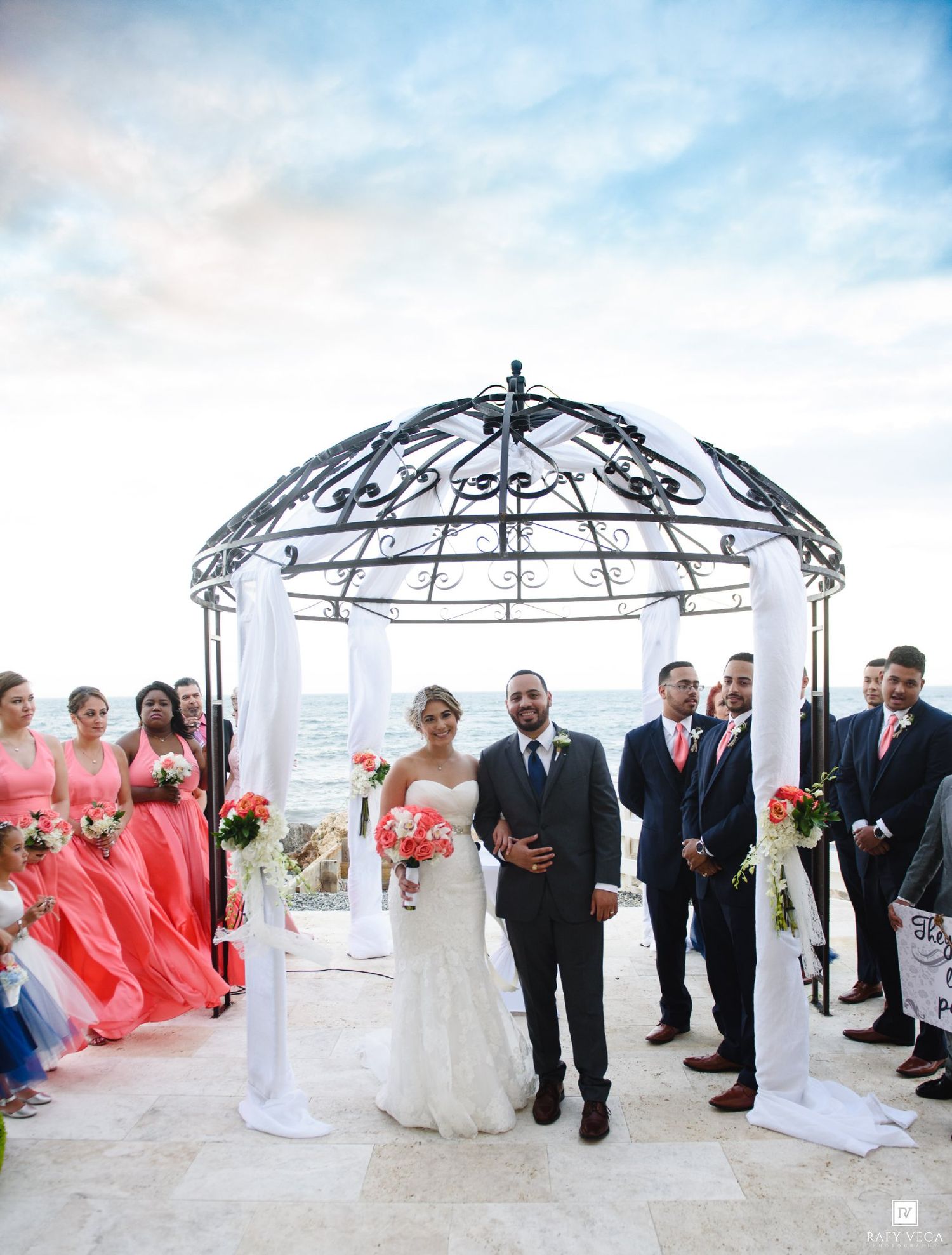 Bride and groom stand under elegant iron gazebo by the ocean, surrounded by coral-dressed bridesmaids and groomsmen.
