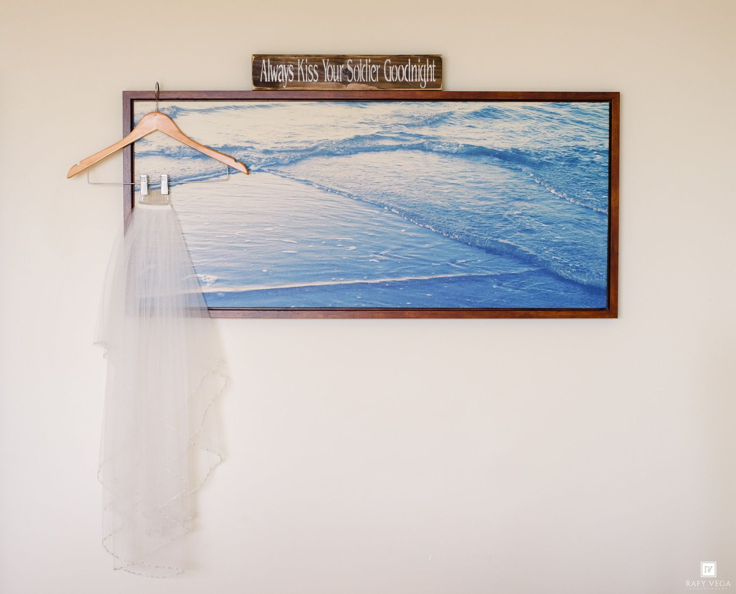 Wedding veil hanging on a wooden hanger in front of a framed ocean pier painting with inspirational quote sign above.