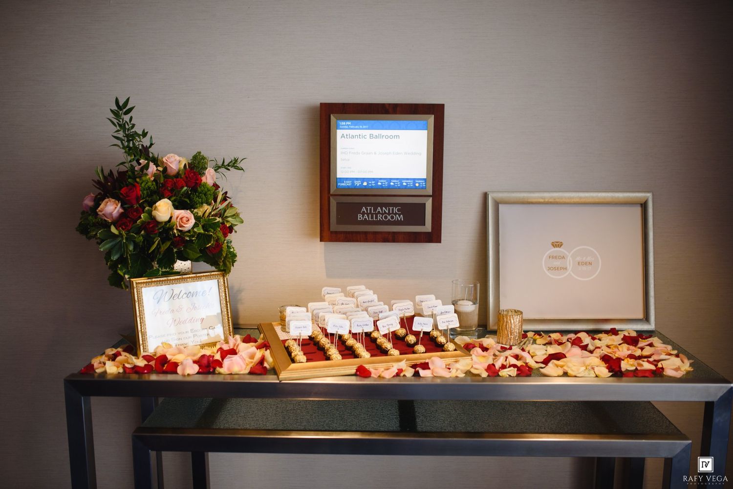 Elegant wedding seating chart display table adorned with rose petals, framed signs, and a floral arrangement.