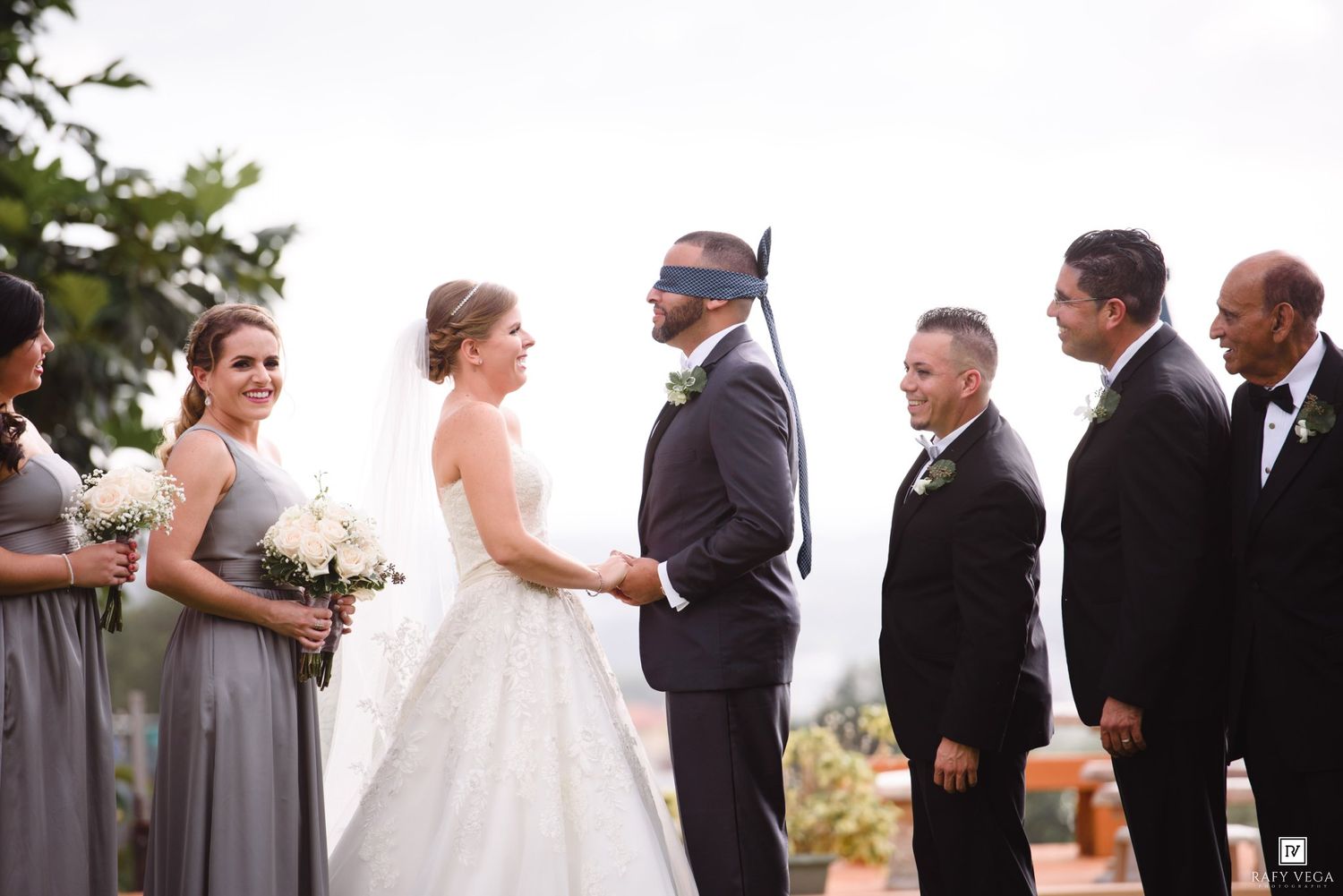 Blindfolded groom holds bride's hands at wedding ceremony with bridesmaids and groomsmen.