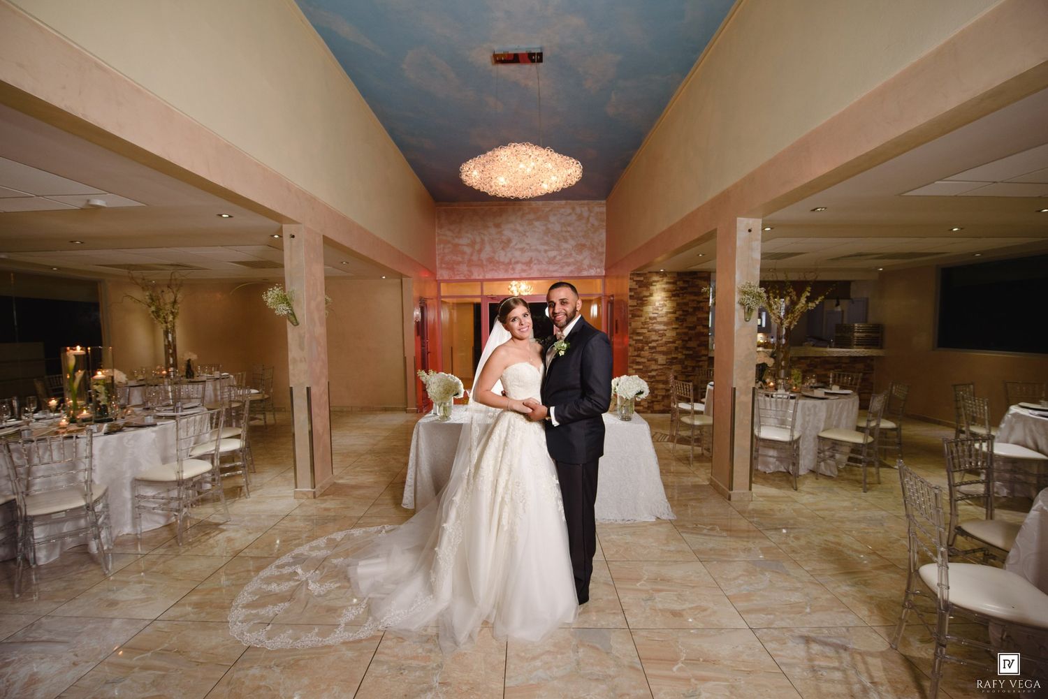 Bride and groom posing elegantly in a decorated wedding reception hall with round tables and a blue ceiling.