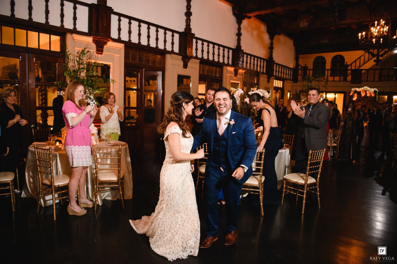 Bride and groom walk joyfully through guests at elegant indoor wedding reception with gold chairs.
