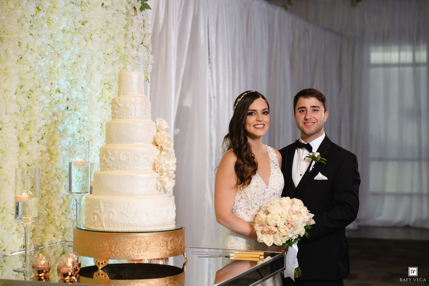 Bride and groom smiling beside a tall white wedding cake at an elegant reception venue.