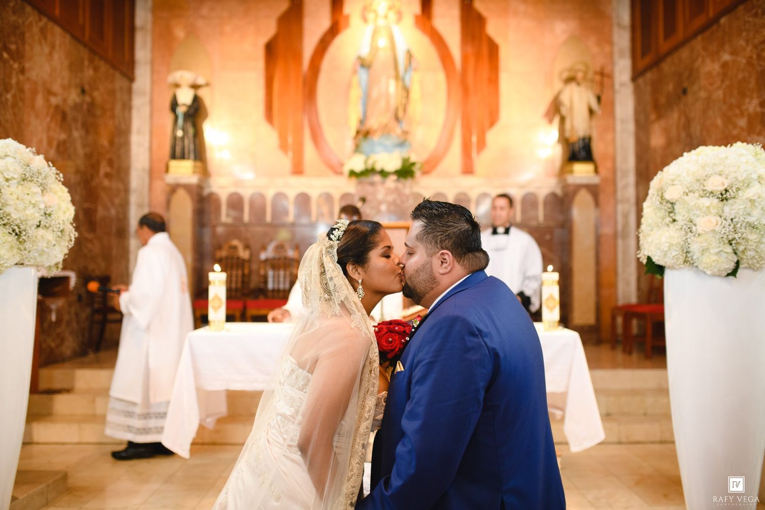 Bride and groom share first kiss at church altar during Catholic wedding ceremony with white floral arrangements.