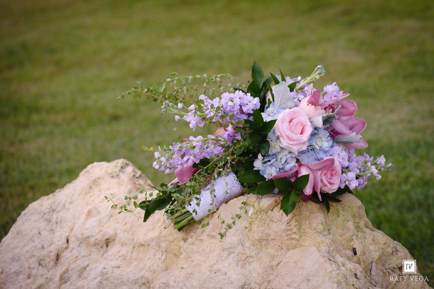 A bridal bouquet of pink roses and purple flowers rests on a large stone with green grass in the background.