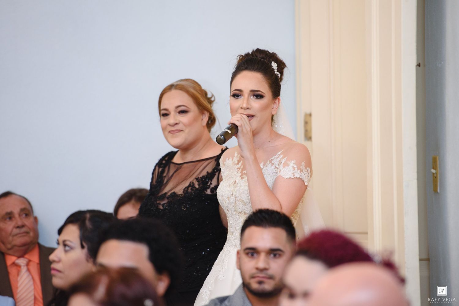 Bride in white lace gown speaking at microphone beside woman in black dress at wedding reception.