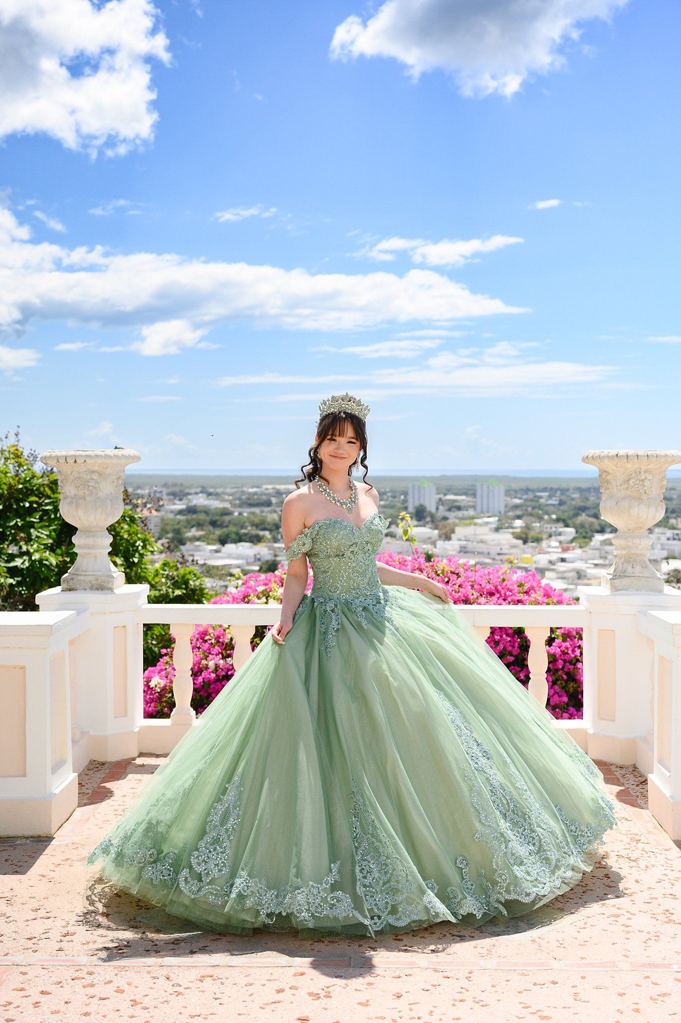 Young woman in elegant sage green quinceañera ball gown with lace details and tiara on a scenic balcony.