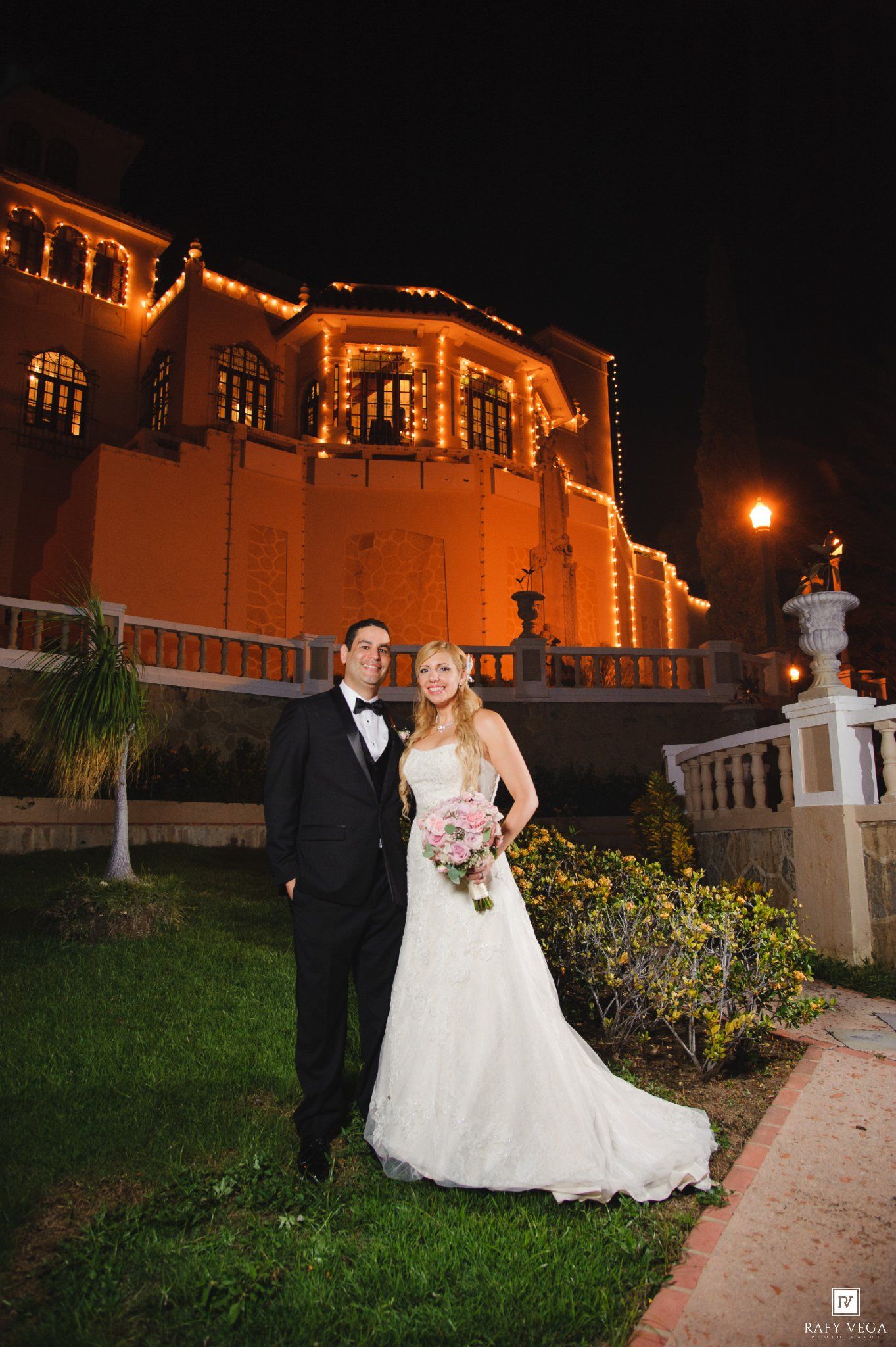 Bride and groom pose outside illuminated wedding venue at night, bride holding pink bouquet in white gown.