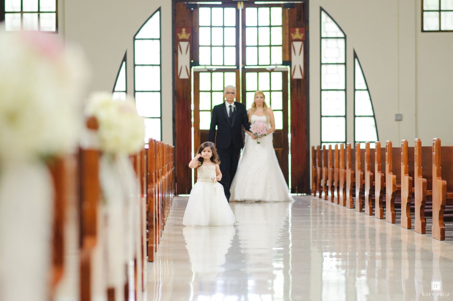 Bride walks down church aisle with flower girl and escort during elegant wedding ceremony.