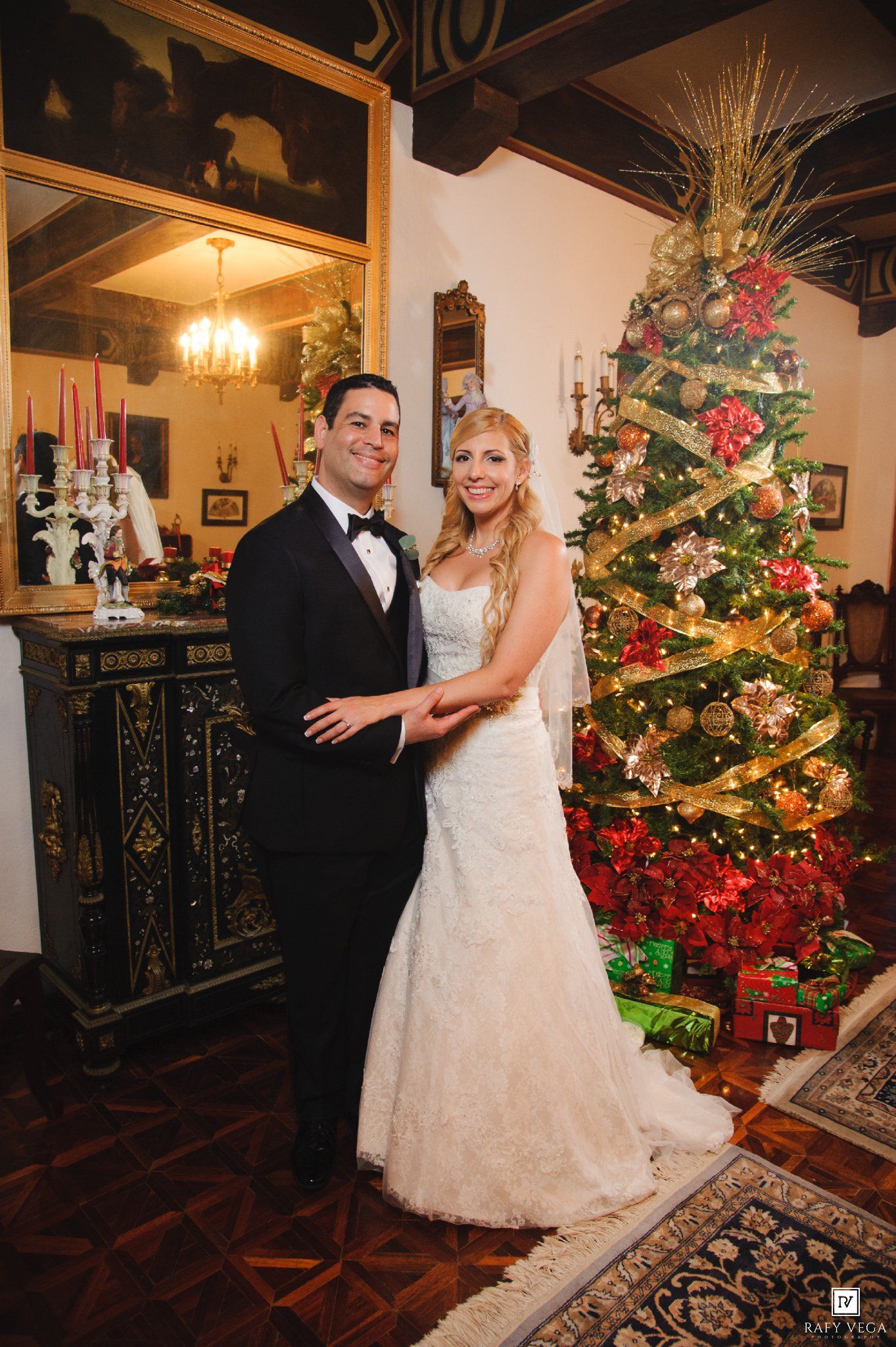 Smiling bride and groom posing together in front of a decorated Christmas tree at their wedding.