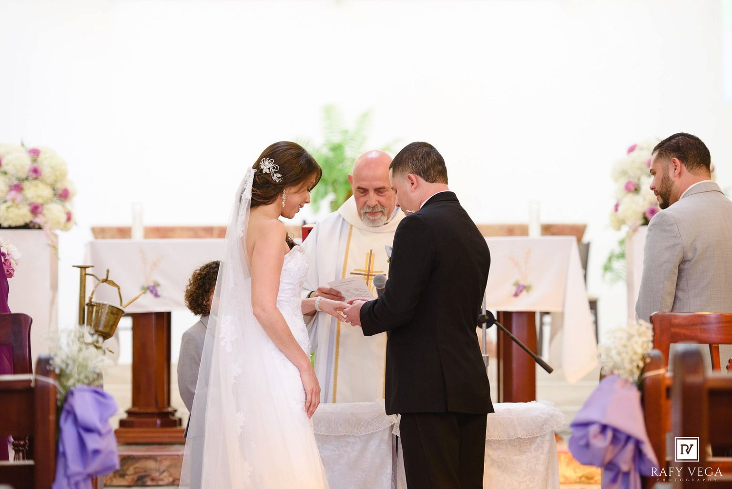 Bride and groom exchanging vows with officiant at a church wedding altar decorated with flowers.