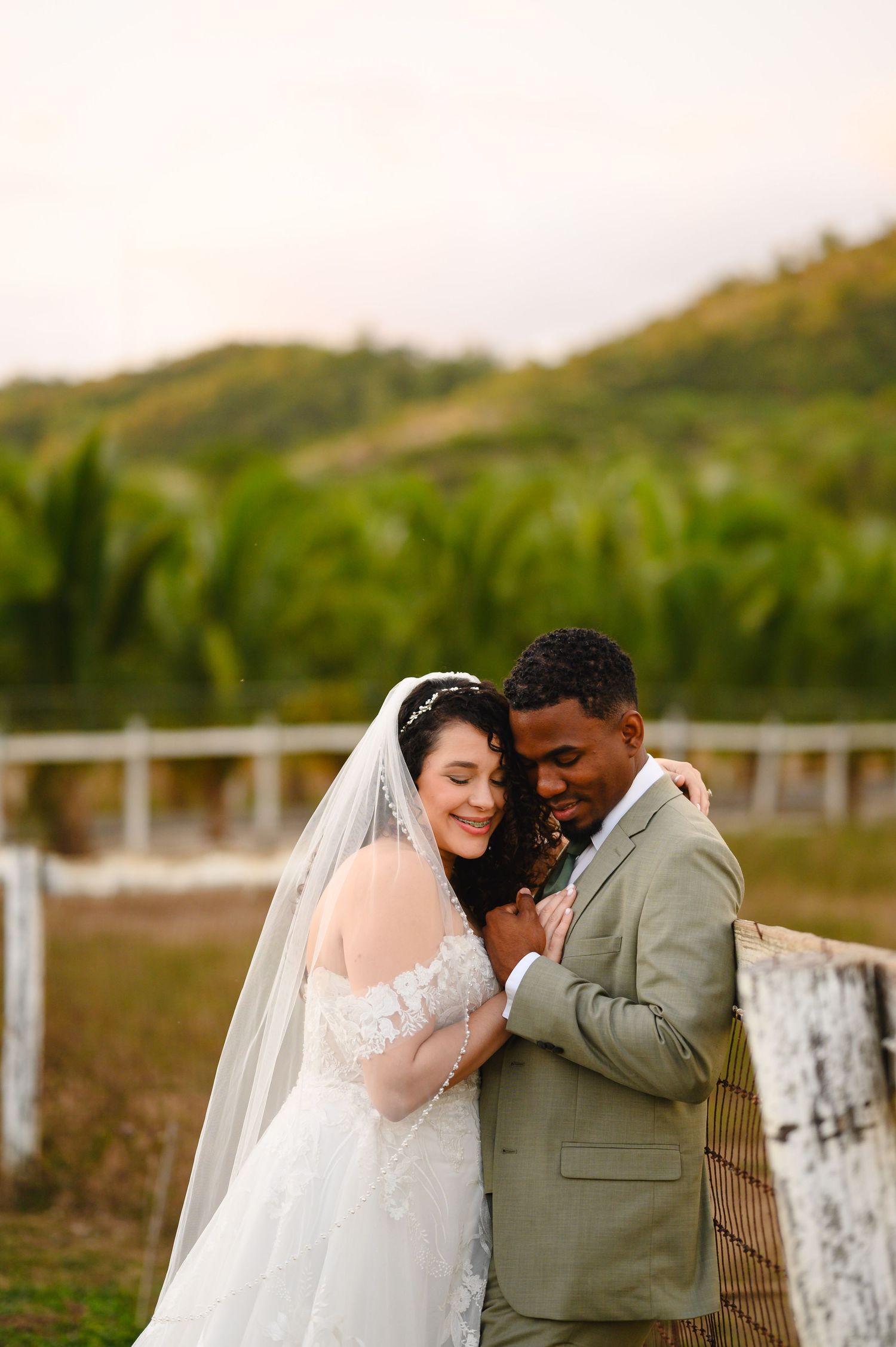 A bride in a lace gown and veil embraces her groom in a sage suit outdoors with lush green hills behind them.