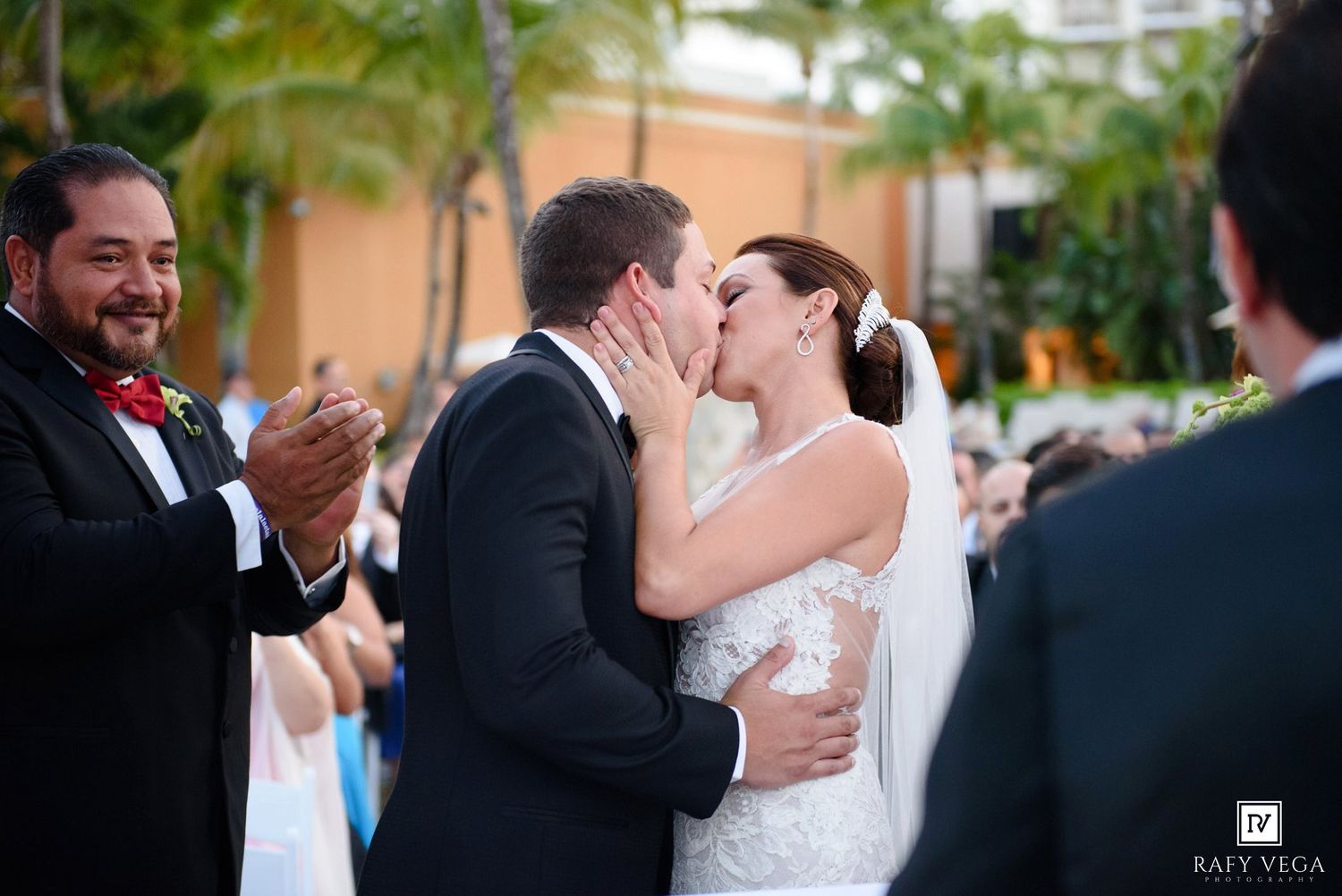 Bride and groom share their first kiss at outdoor wedding ceremony while guests applaud in tropical venue.