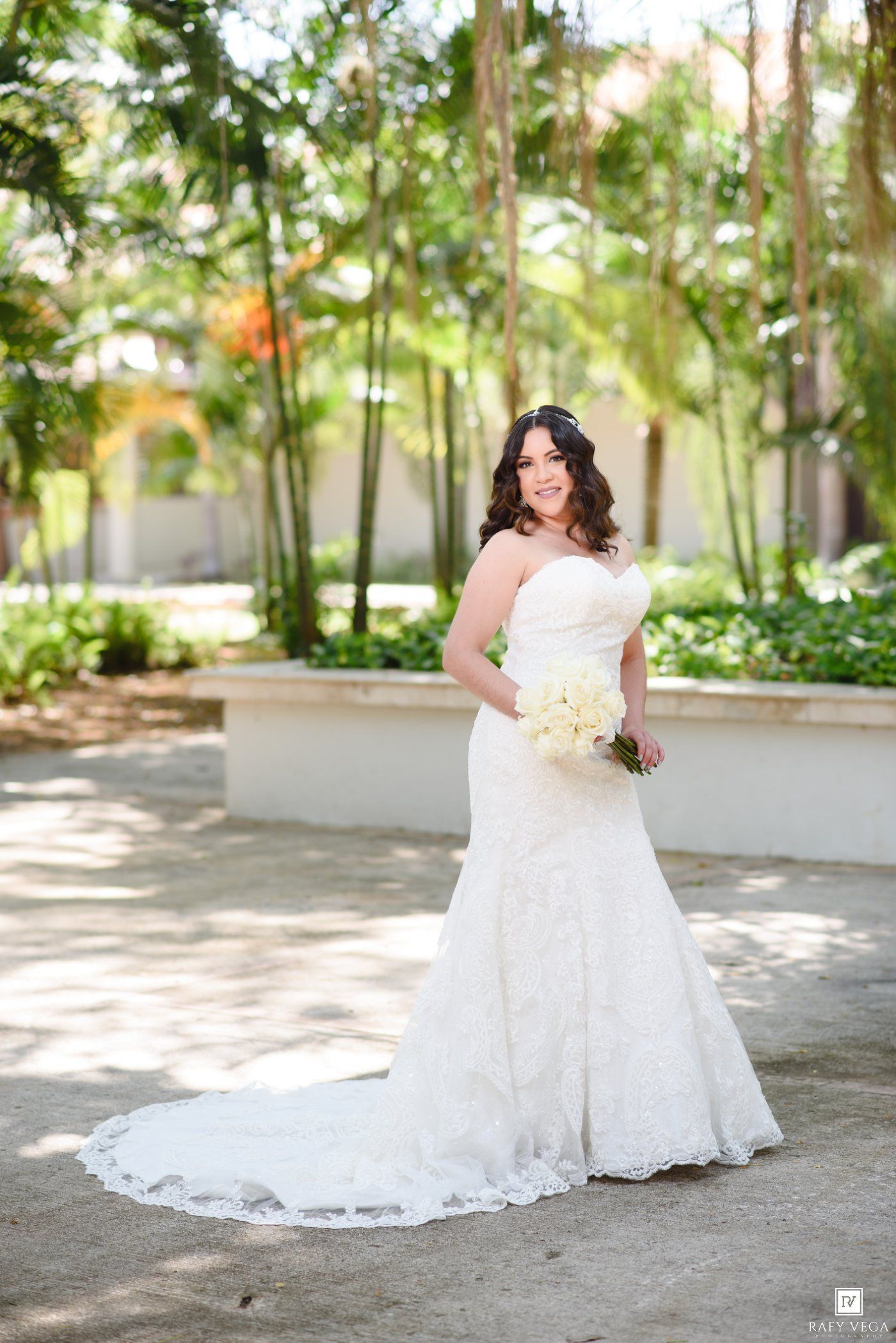 Bride in strapless lace mermaid wedding gown holding white bouquet poses outdoors among lush green trees.