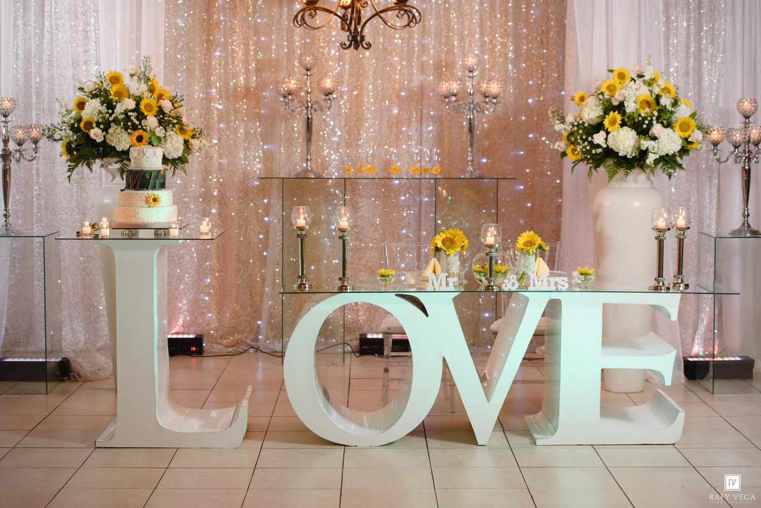 Wedding reception decor featuring large LOVE letters, white cake, yellow and white floral arrangements with fairy lights.