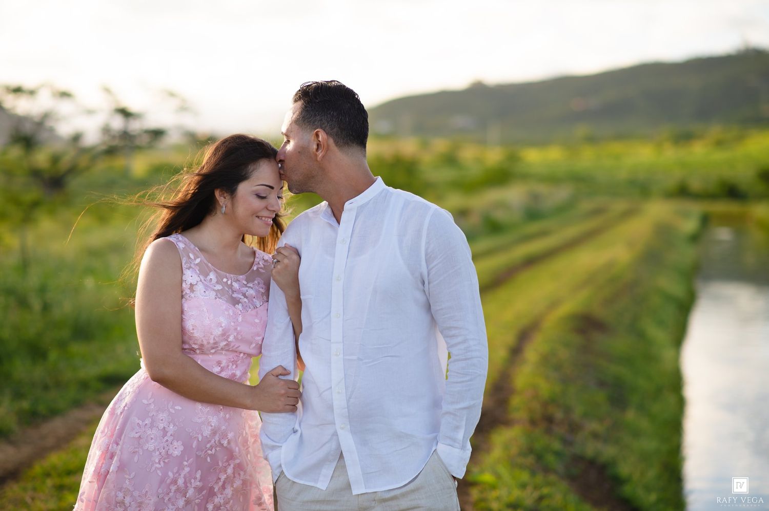 Couple sharing a romantic moment in a lush green field at golden hour, woman in pink dress, man in white shirt.