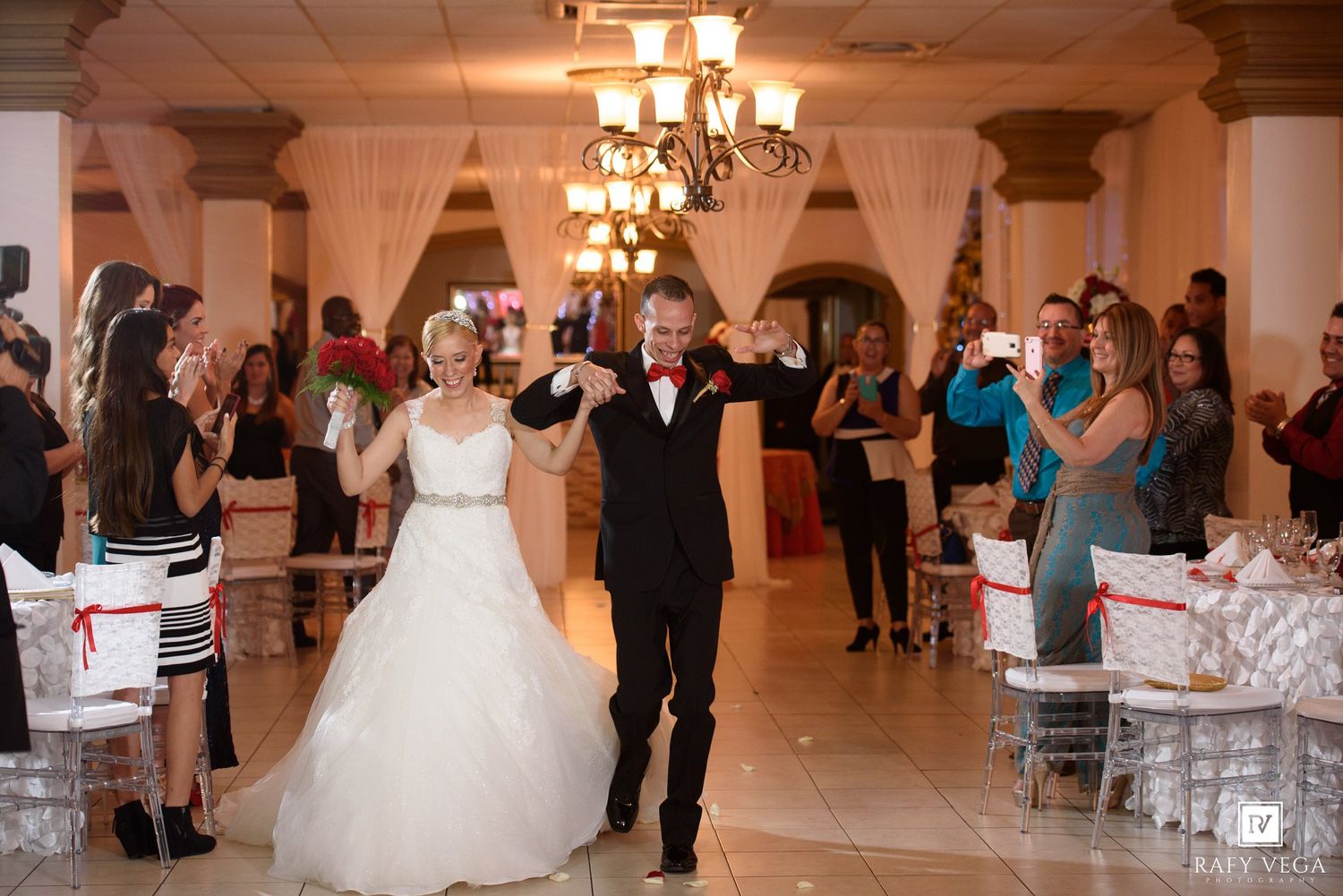 Bride and groom make grand entrance at elegant wedding reception with guests cheering in decorated ballroom.