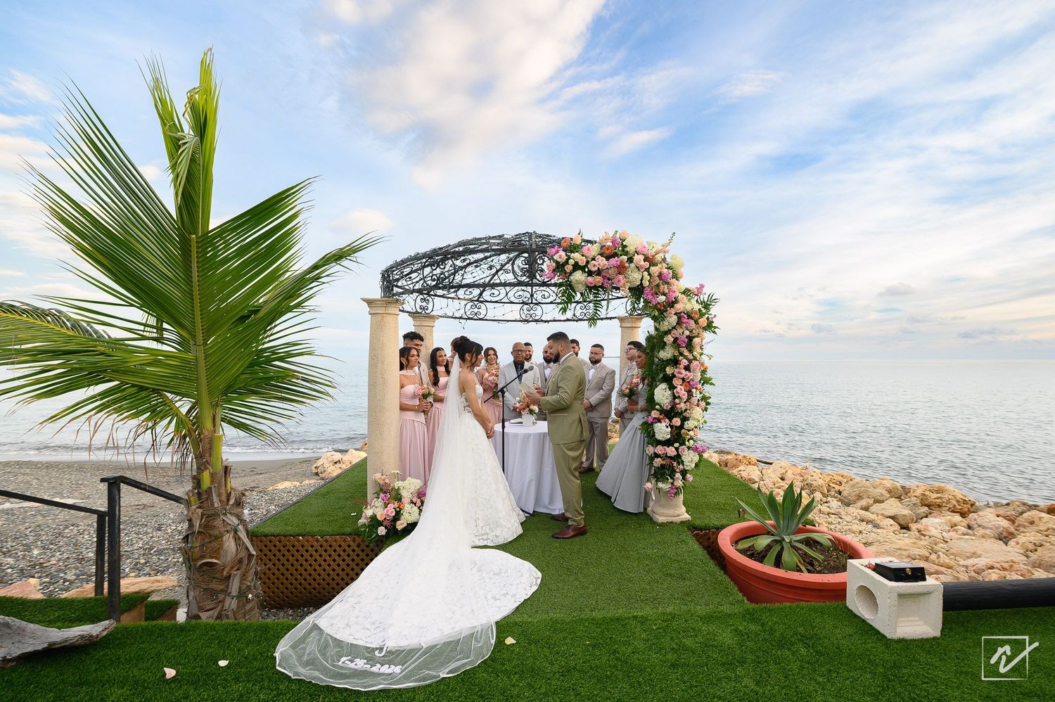 Bride in a long white gown and groom exchange vows under a floral-covered gazebo on a green lawn beside the ocean at a beachfront wedding.