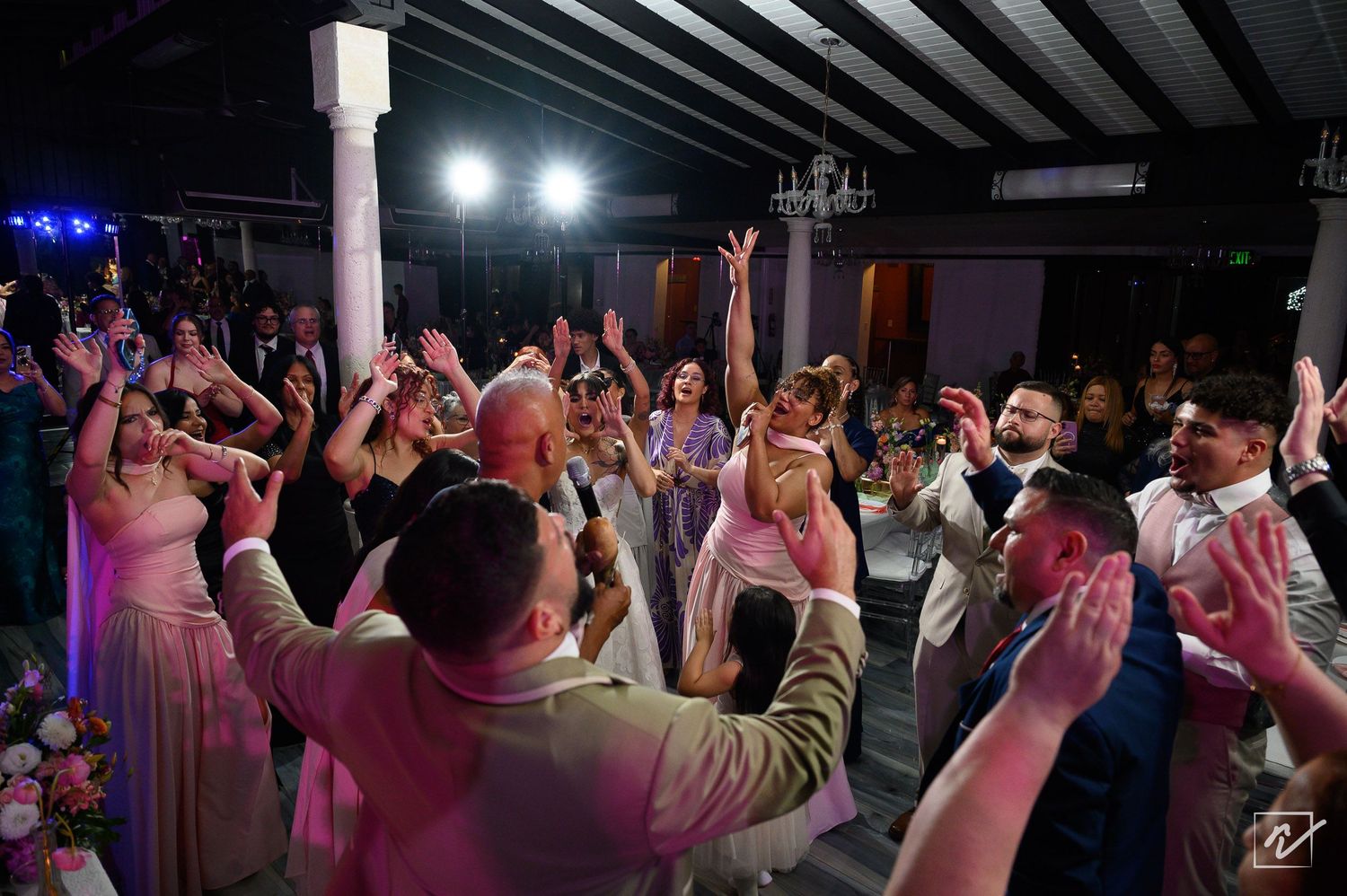 Wedding guests raise their hands and cheer in a lively circle on the dance floor during an energetic evening reception.